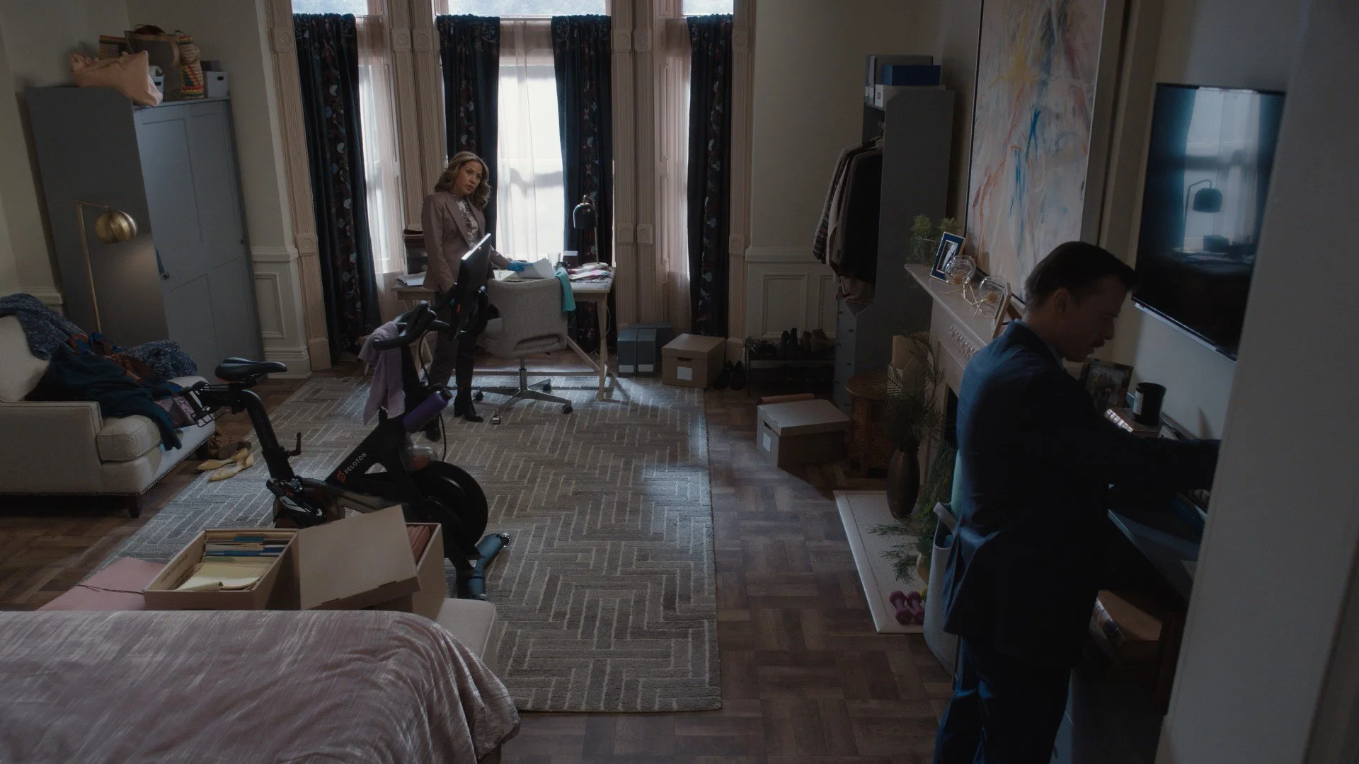 A living room with a woman standing near a desk, and a man in a suit sitting at a piano. The room has large windows with dark curtains, a gray sofa with clothes on it, an exercise bike, and various boxes and books.