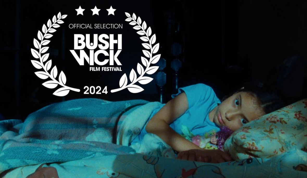 A young girl lying on her side in bed, resting her head on a floral pillow, with a faint smile, wearing a light blue shirt, in a dimly lit room.