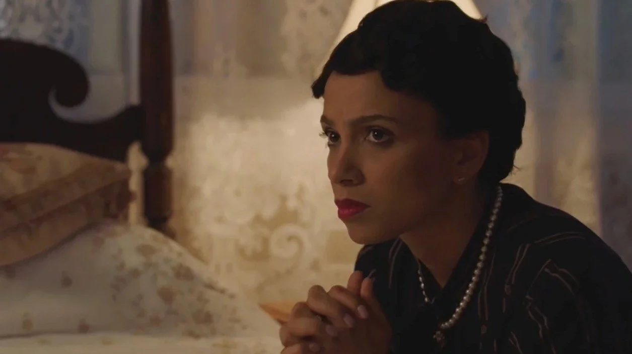 A woman with short dark hair, wearing a pearl necklace, sits at a table looking to her right with a serious expression inside a dimly lit room.