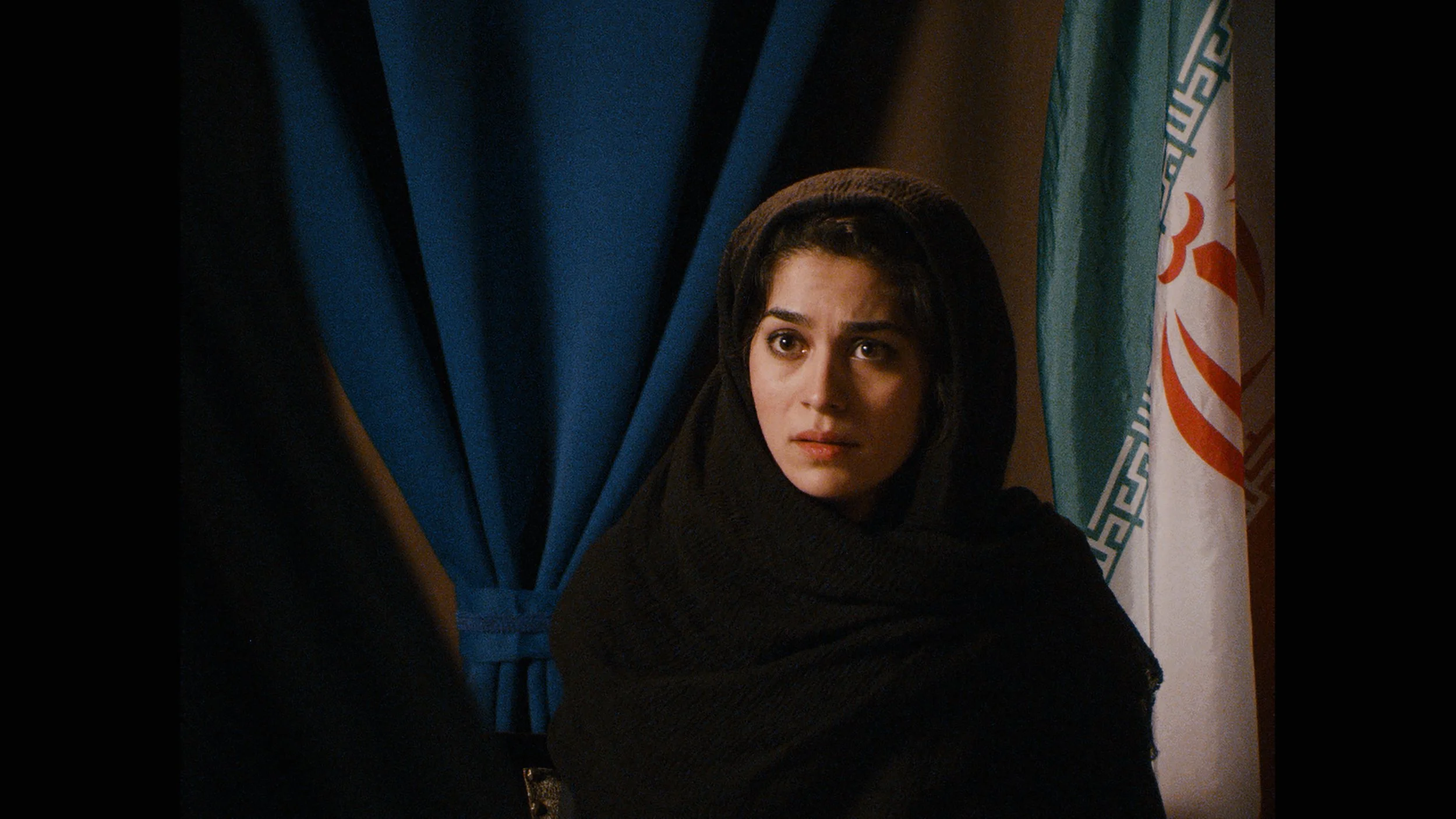 A woman wearing a black headscarf sitting in front of blue curtains and a flag with Persian script.