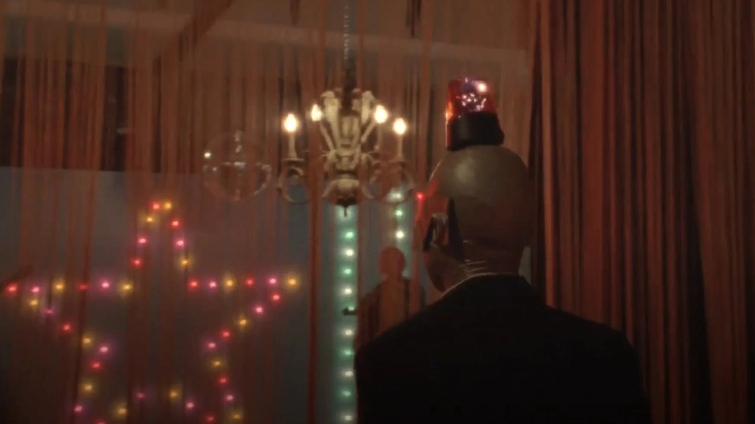 A scene from a film showing a person dressed as a robot or robot-like figure with a metallic head, looking at a circus or carnival mirror with colorful lights and a chandelier in the background.