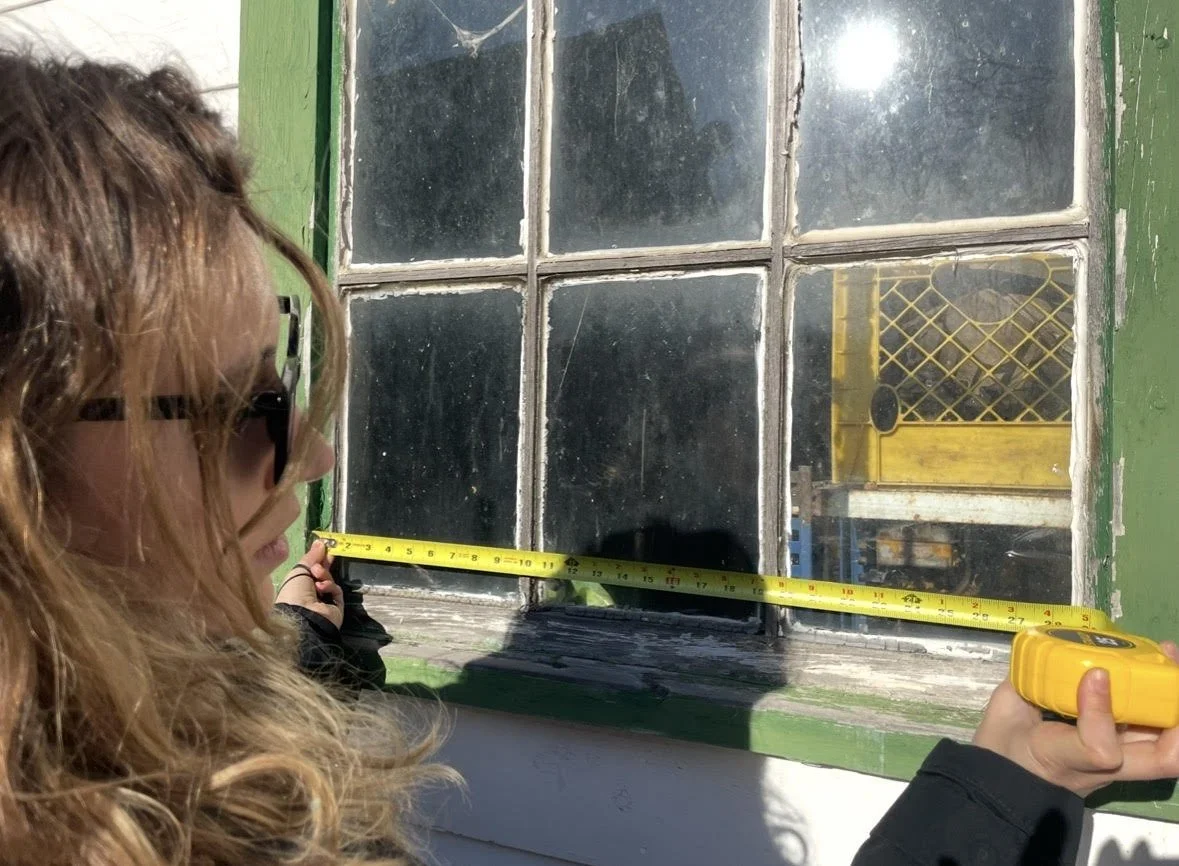 A woman measuring a wooden window frame with a tape measure inside a building.