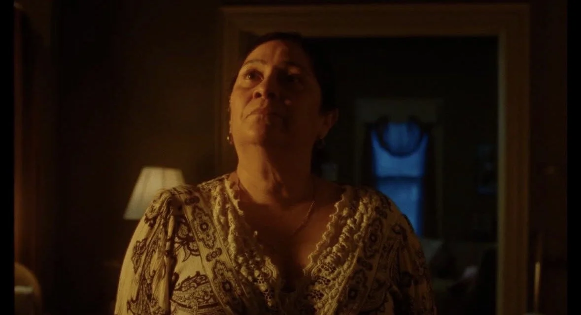 A woman standing in a dimly lit room with a serious expression, wearing a patterned blouse, with a lamp and a window with curtains in the background.