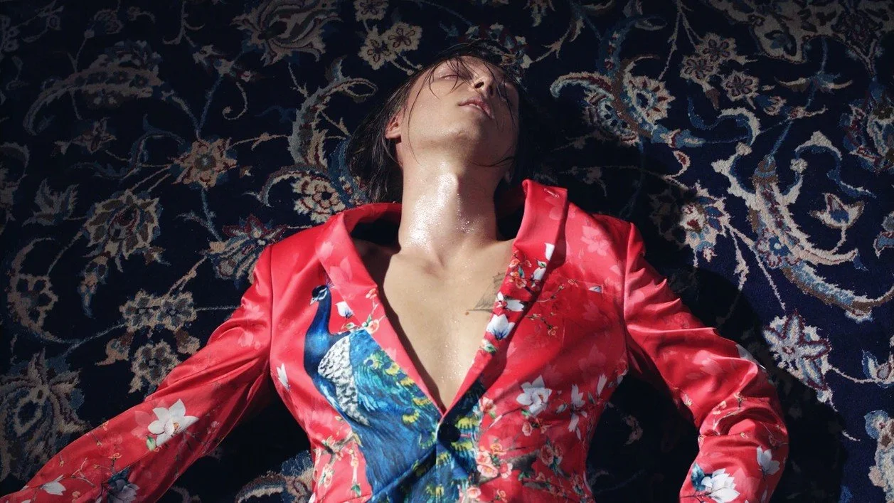 A person lying on a patterned carpet, wearing a red silk shirt with a peacock design, with their eyes closed and face tilted upward.