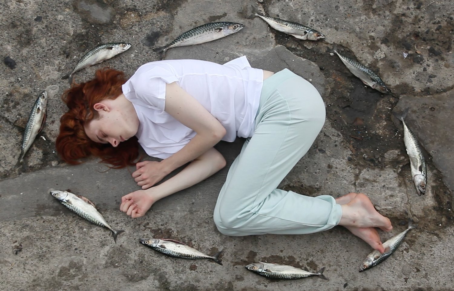 A woman with red hair lying on her side on a rocky and sandy surface, surrounded by dead fish.