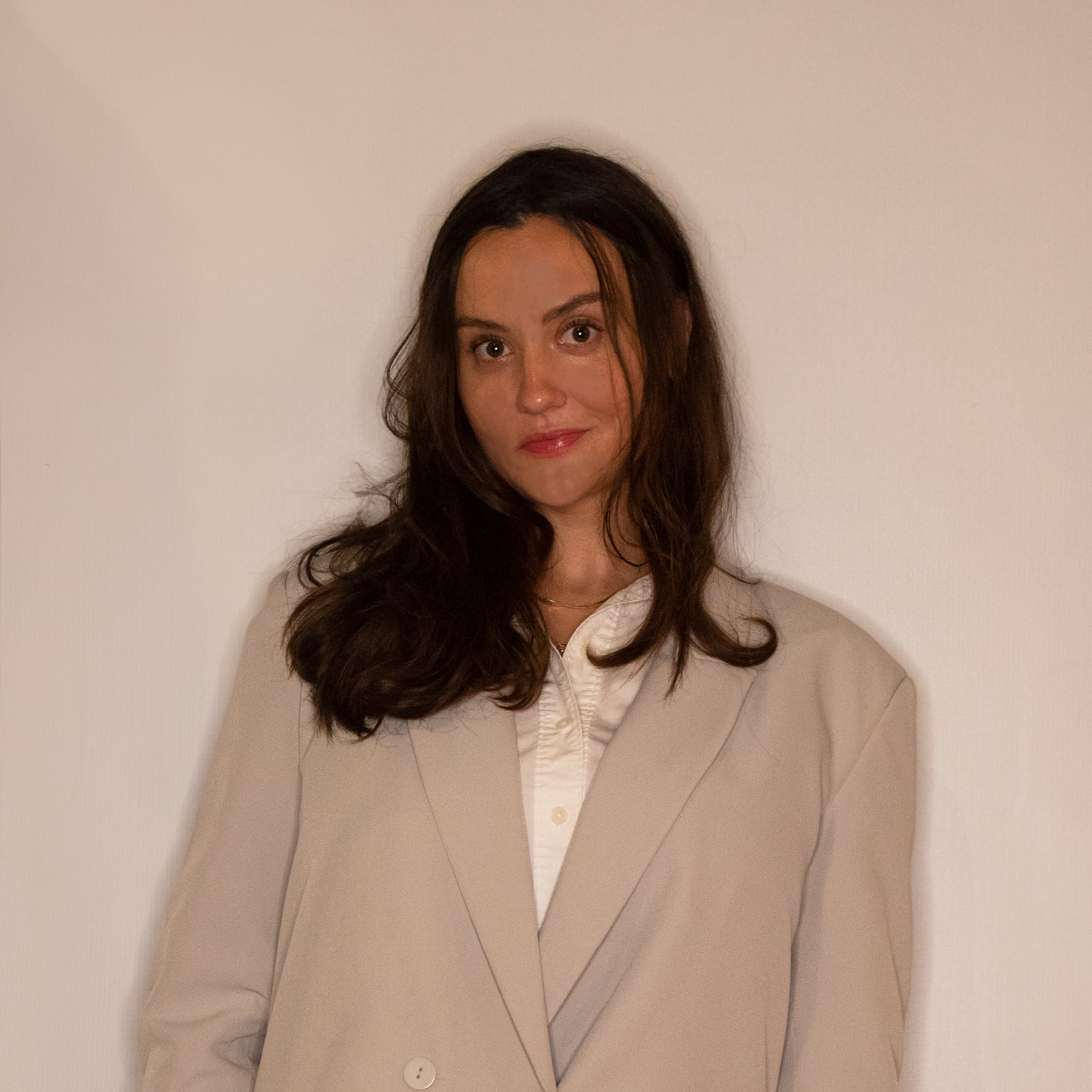 A woman with long dark hair wearing a beige blazer and a white blouse standing against a plain white background.
