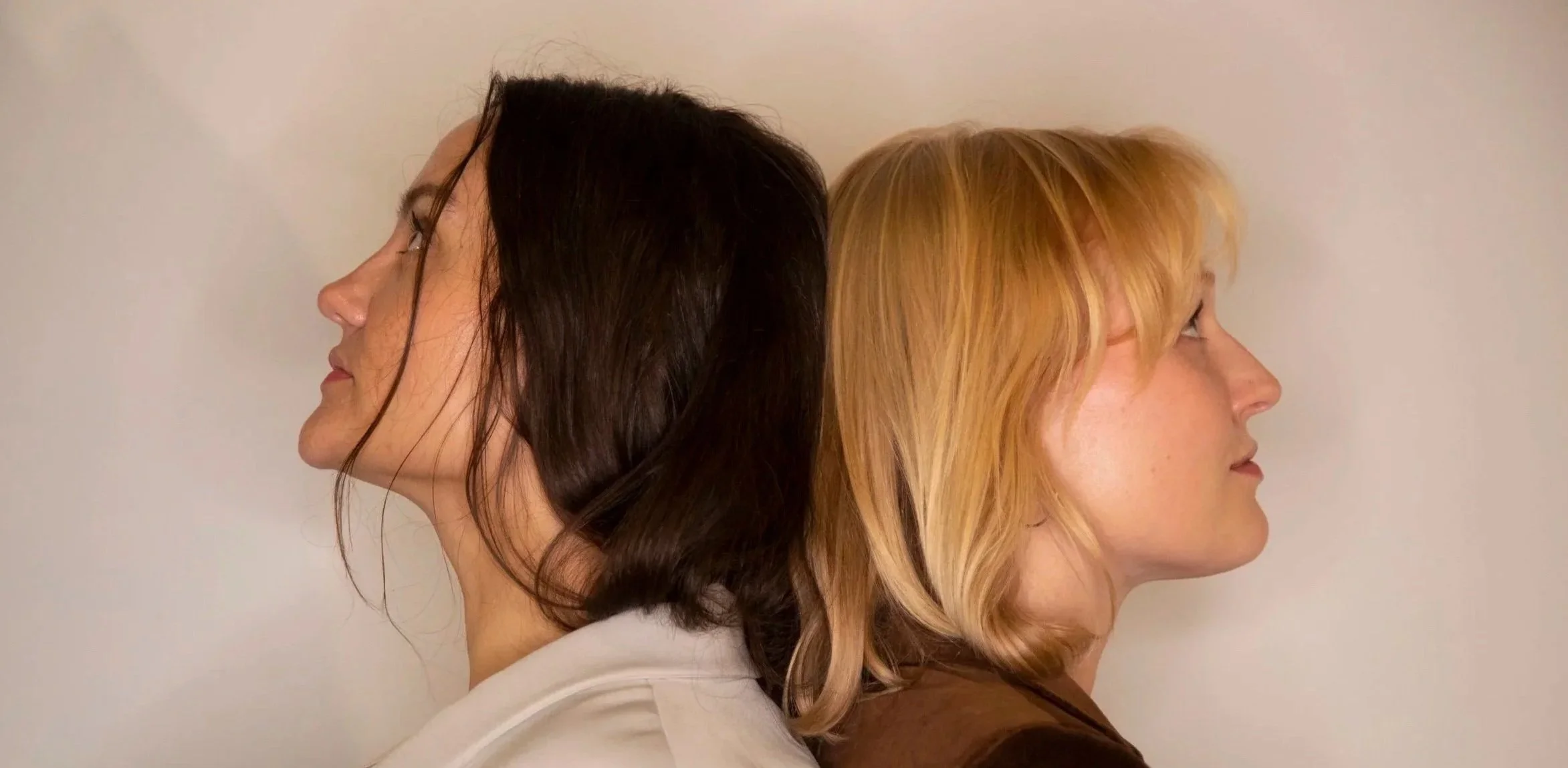 Two women standing back-to-back with faces in profile, one with dark hair and the other with blonde hair, against a plain background.