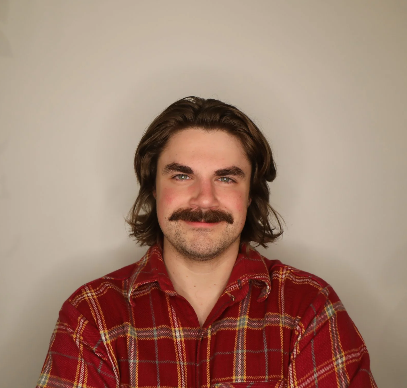 Portrait of a man with long wavy hair, a mustache, wearing a red plaid shirt, smiling, against a plain light-colored background.