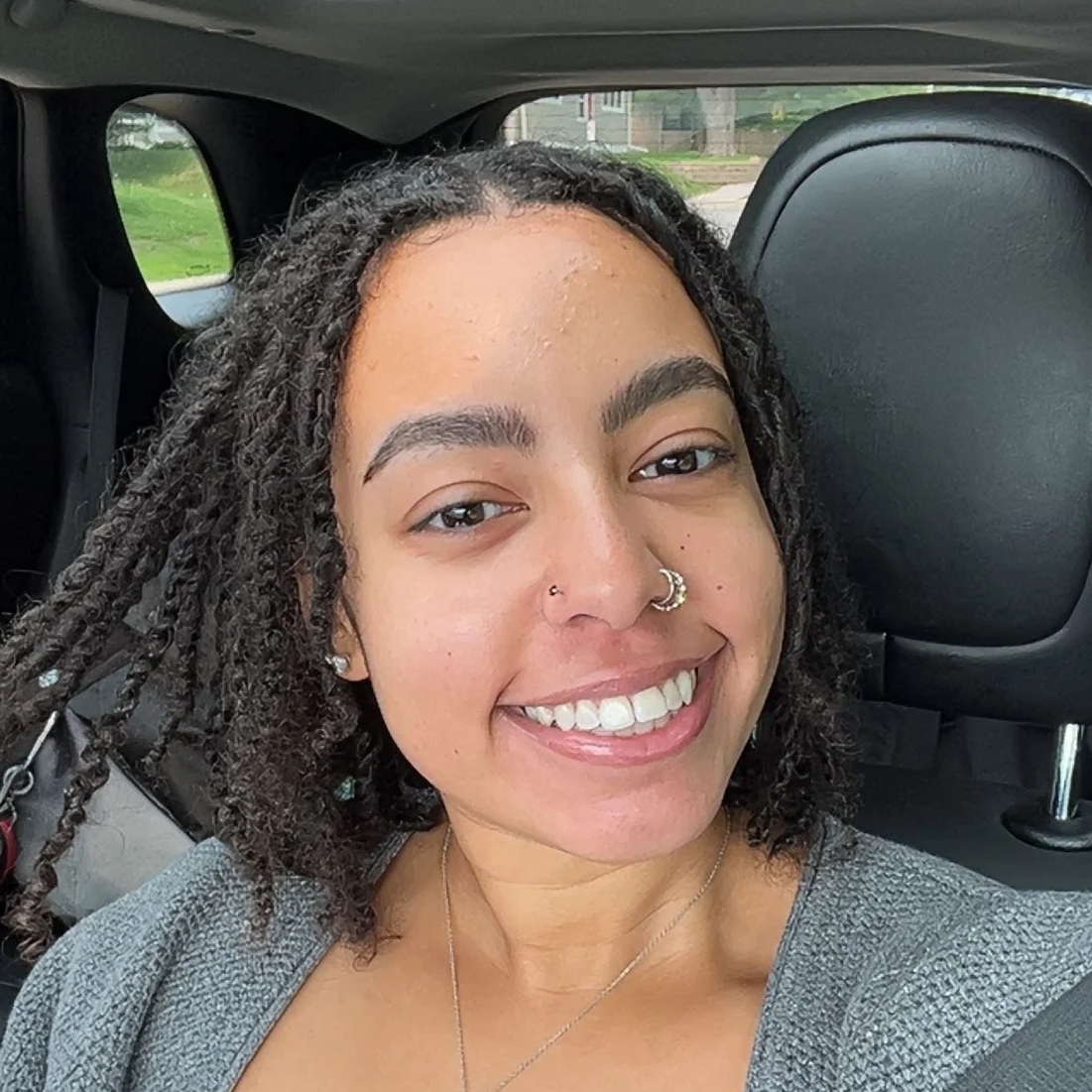 Smiling woman with curly hair, wearing a gray top, sitting in a car.