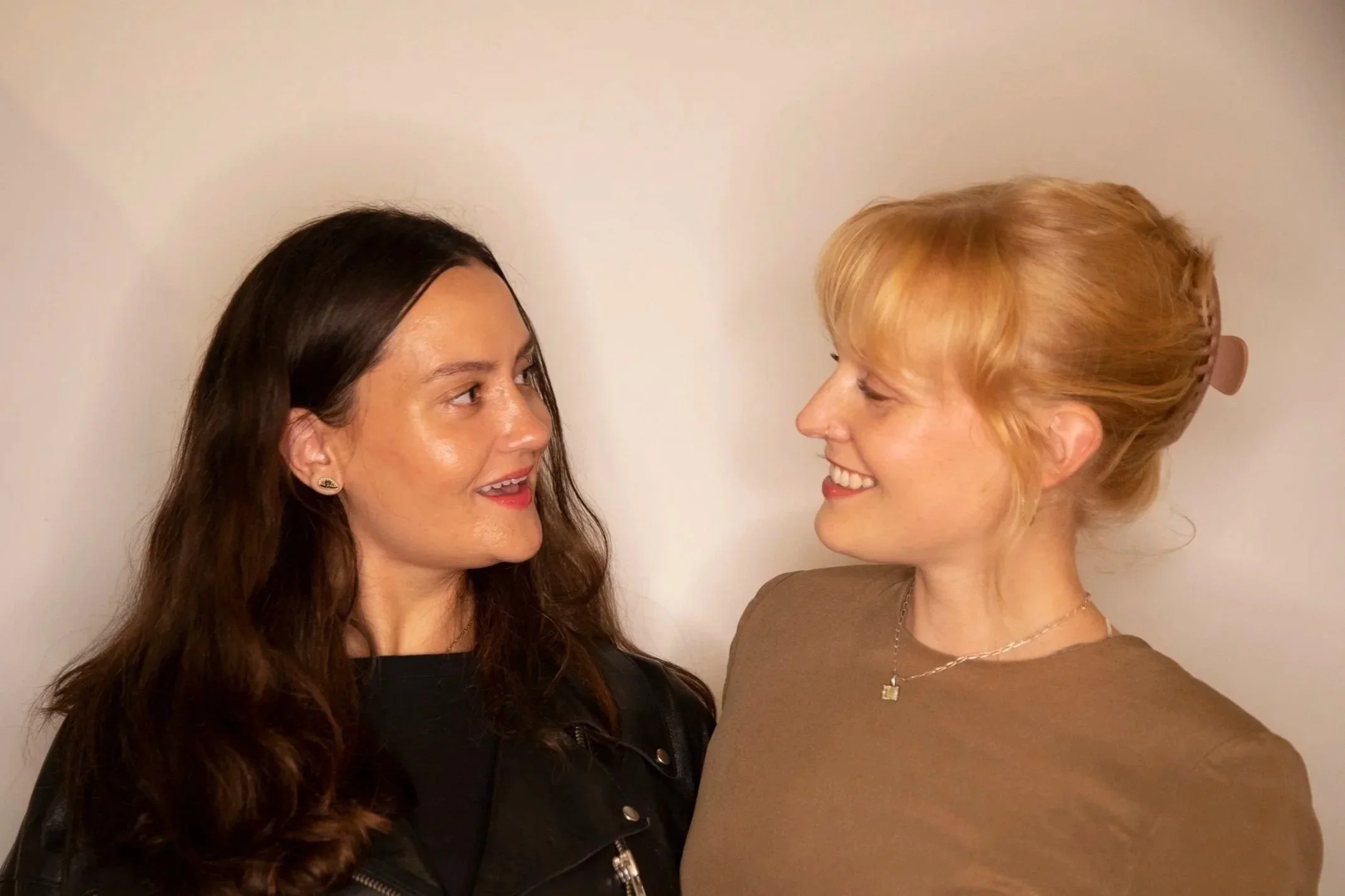 Two women face each other, smiling and looking into each other's eyes against a plain background.