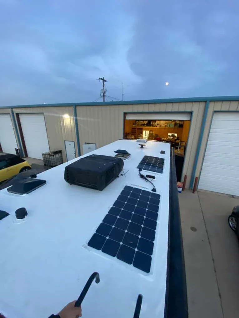 "Resurfaced RV roof with resealed vents, skylights, and mounted solar panels outside a repair shop bay."