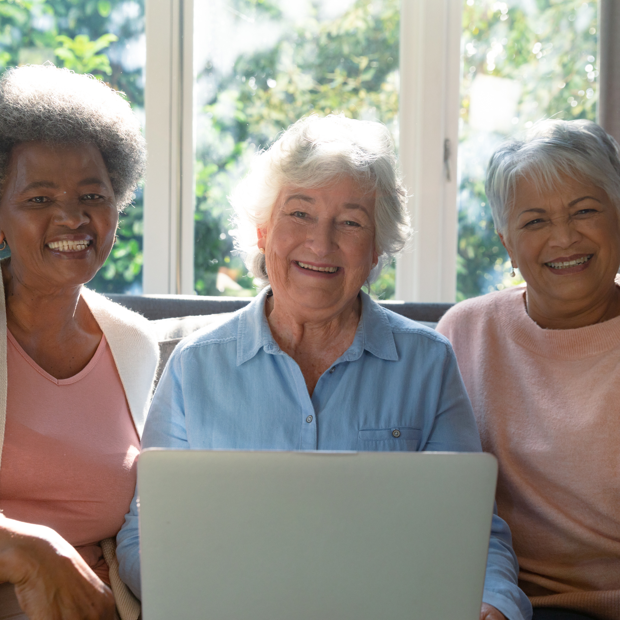 Three smiling elderly women sitting on a couch in front of a window, looking at a laptop.