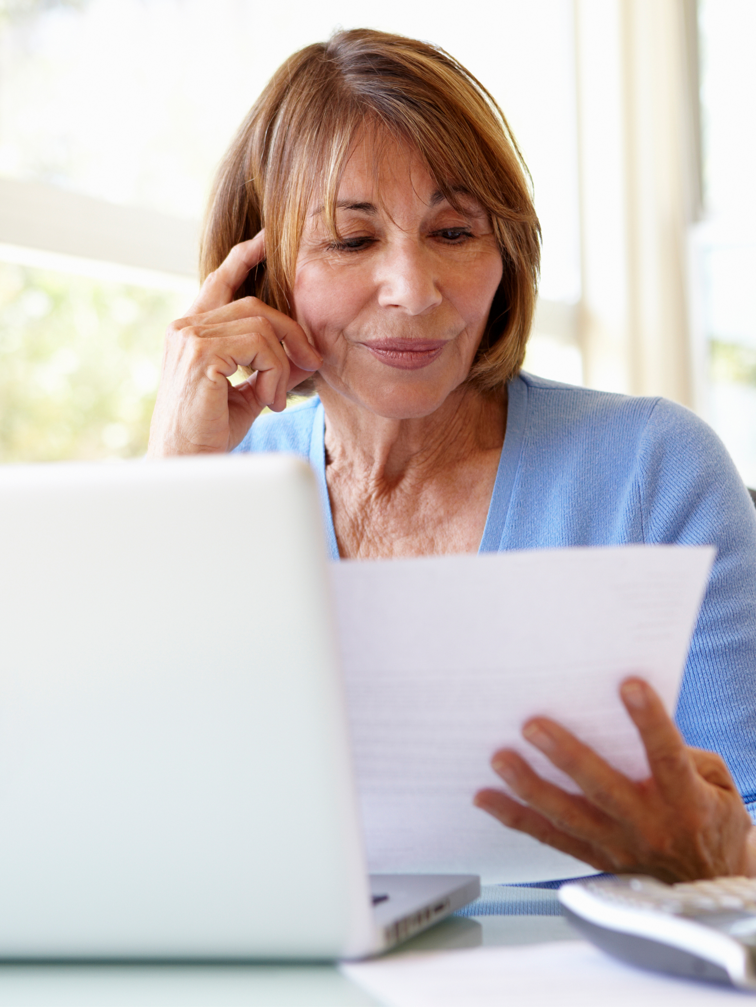 A woman looking at a piece of paper with a laptop in front of her, sitting at a desk in a well-lit room.