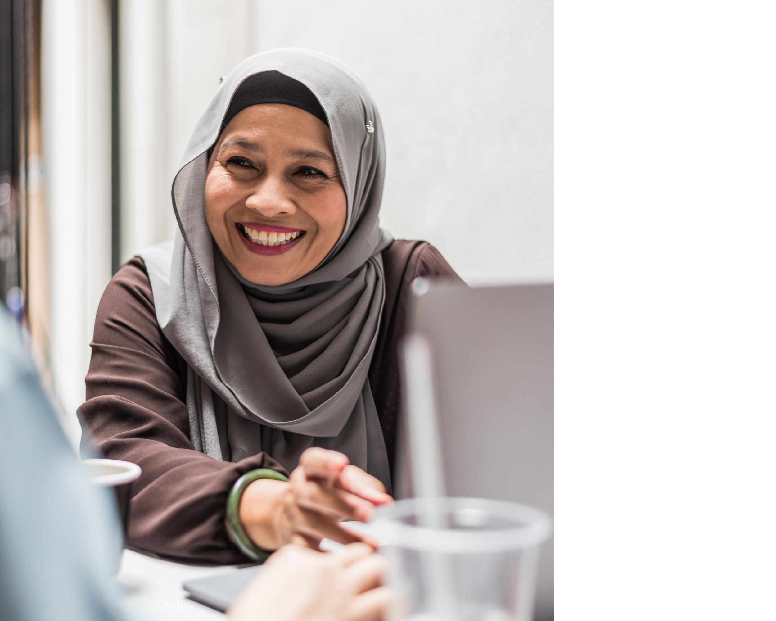 A woman wearing a gray hijab and brown top smiling while working on a laptop.