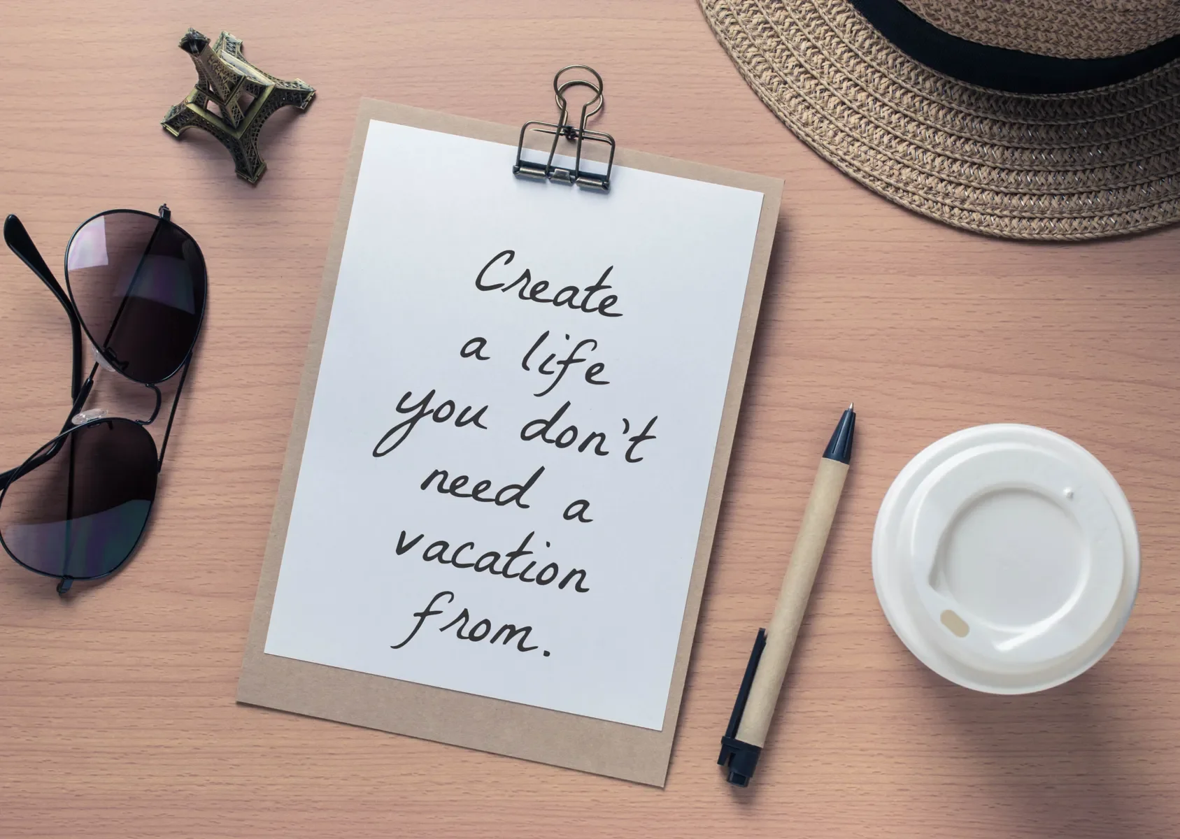 A wooden desk with a paper clip holding a white paper that has the quote 'Create a life you don't need a vacation from.' A pen, a disposable coffee cup lid, sunglasses, a small Eiffel Tower figurine, and a straw hat are also on the desk.