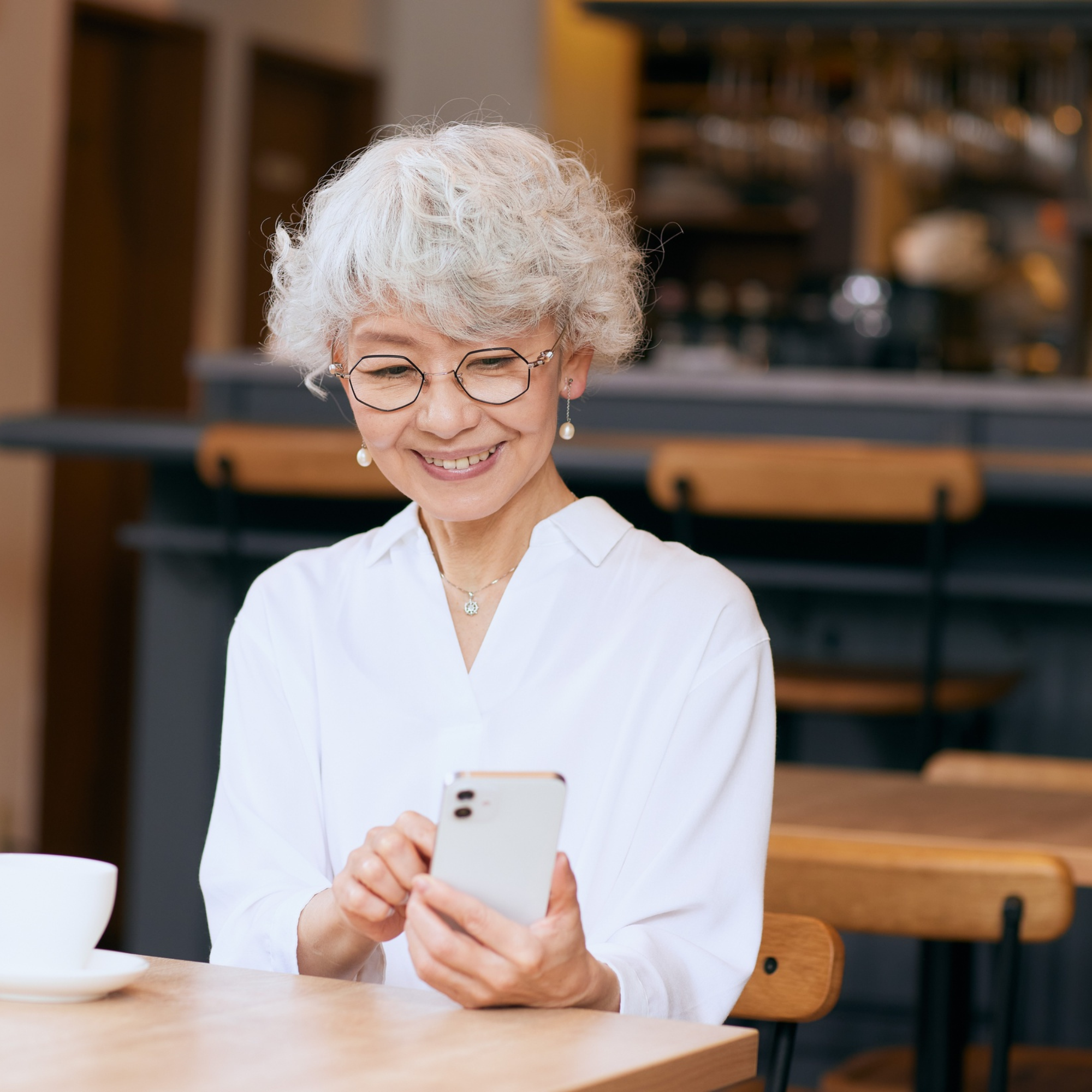 A cheerful elderly woman with gray curly hair and glasses sitting at a wooden table in a cafe, looking at her smartphone and smiling.
