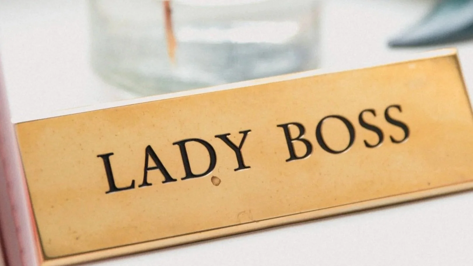 Close-up of a sign that reads 'LADY BOSS' in black letters on a gold background, with a glass object in the blurred background.