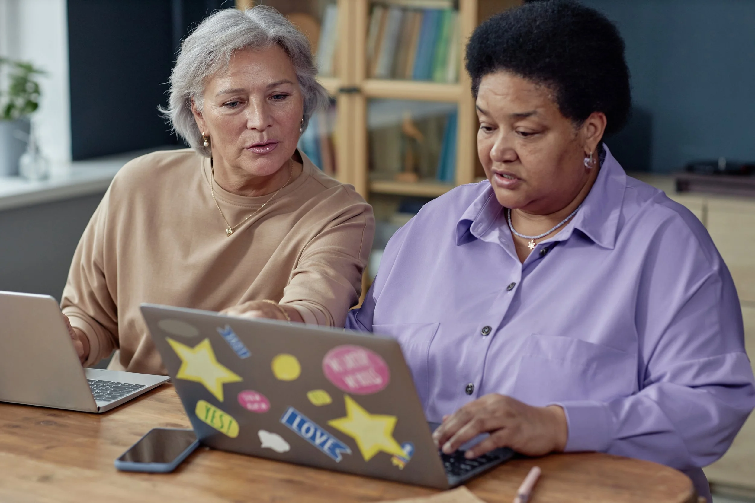 Two women sitting at a wooden table looking at a laptop decorated with stickers, including stars, speech bubbles, and words like "LOVE" and "YES." They are inside a room with a bookshelf and window in the background.