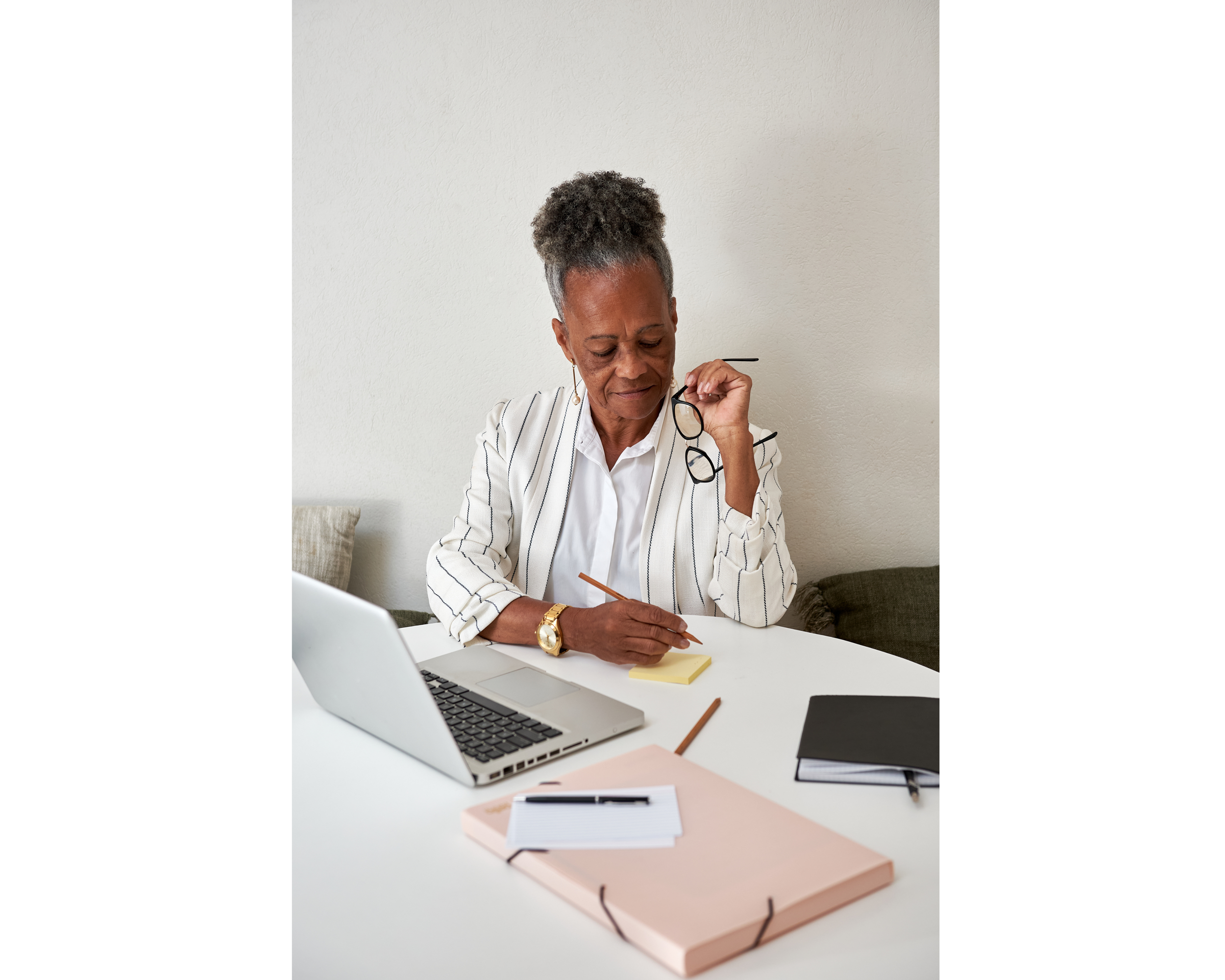 An older woman sitting at a white desk, holding her glasses with one hand and writing on a yellow sticky note with the other, surrounded by a laptop, notebooks, and pens.