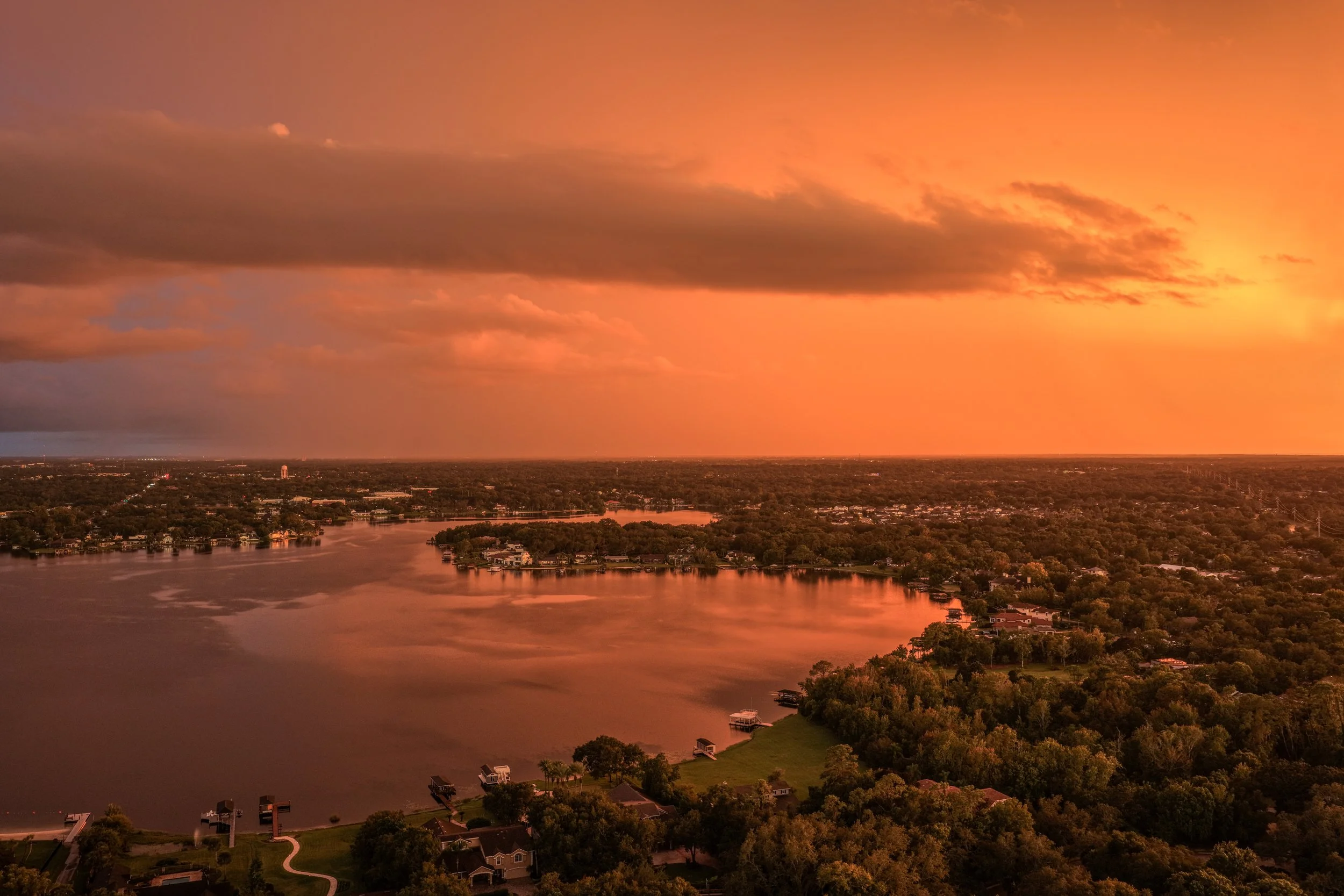Aerial view of a river at sunset with orange sky and clouds, surrounding greenery, and houses near the water.