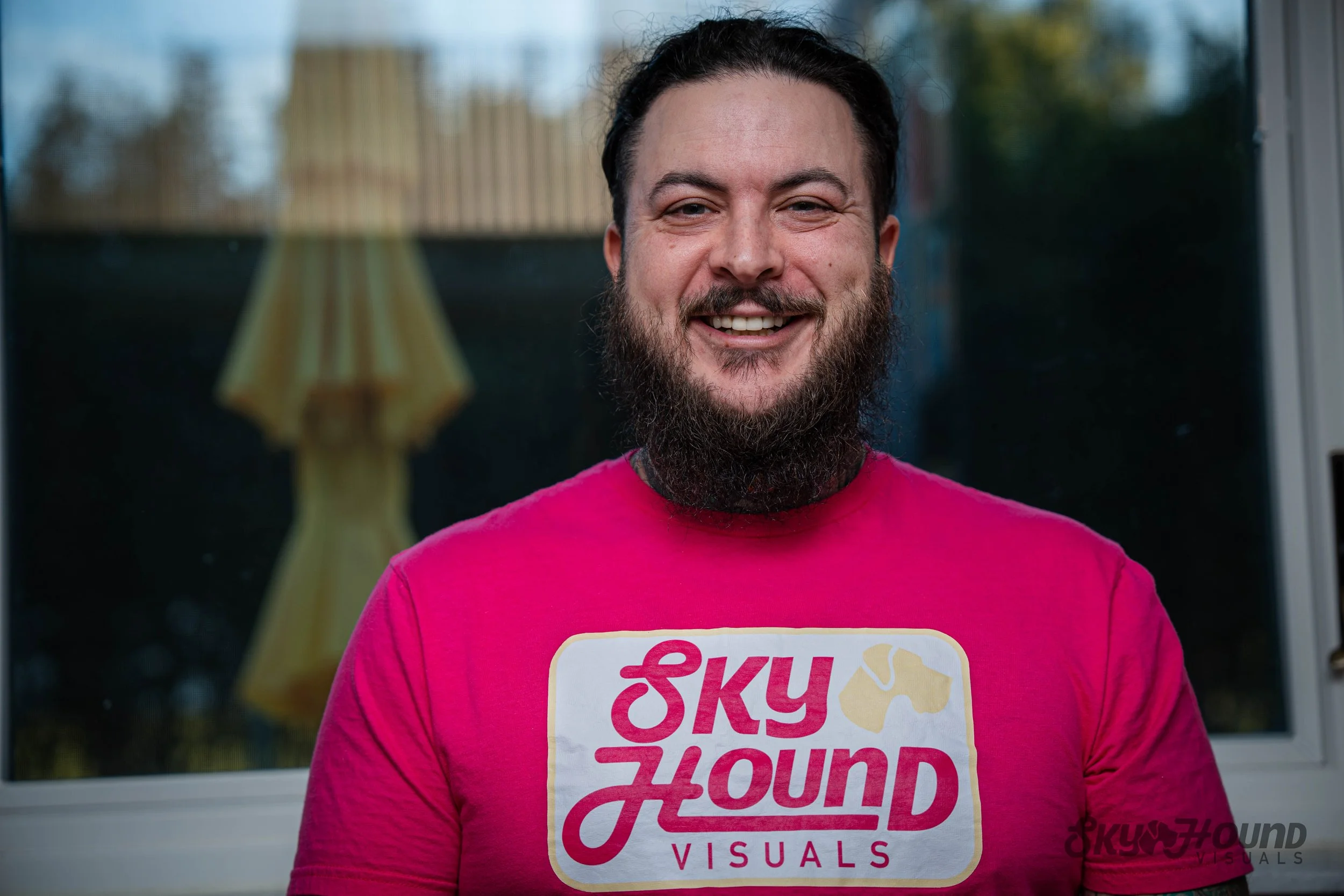 A smiling man with a beard wearing a pink T-shirt that reads 'Sky Hound Visuals' standing in front of a window with a blurred view of trees and a yellow umbrella outside.