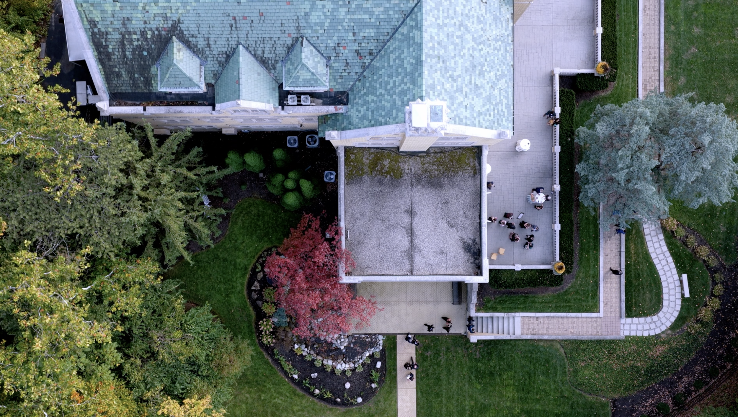 An aerial view of a building with a green, tiled roof, surrounded by landscaped gardens and a paved patio area with some people gathered.