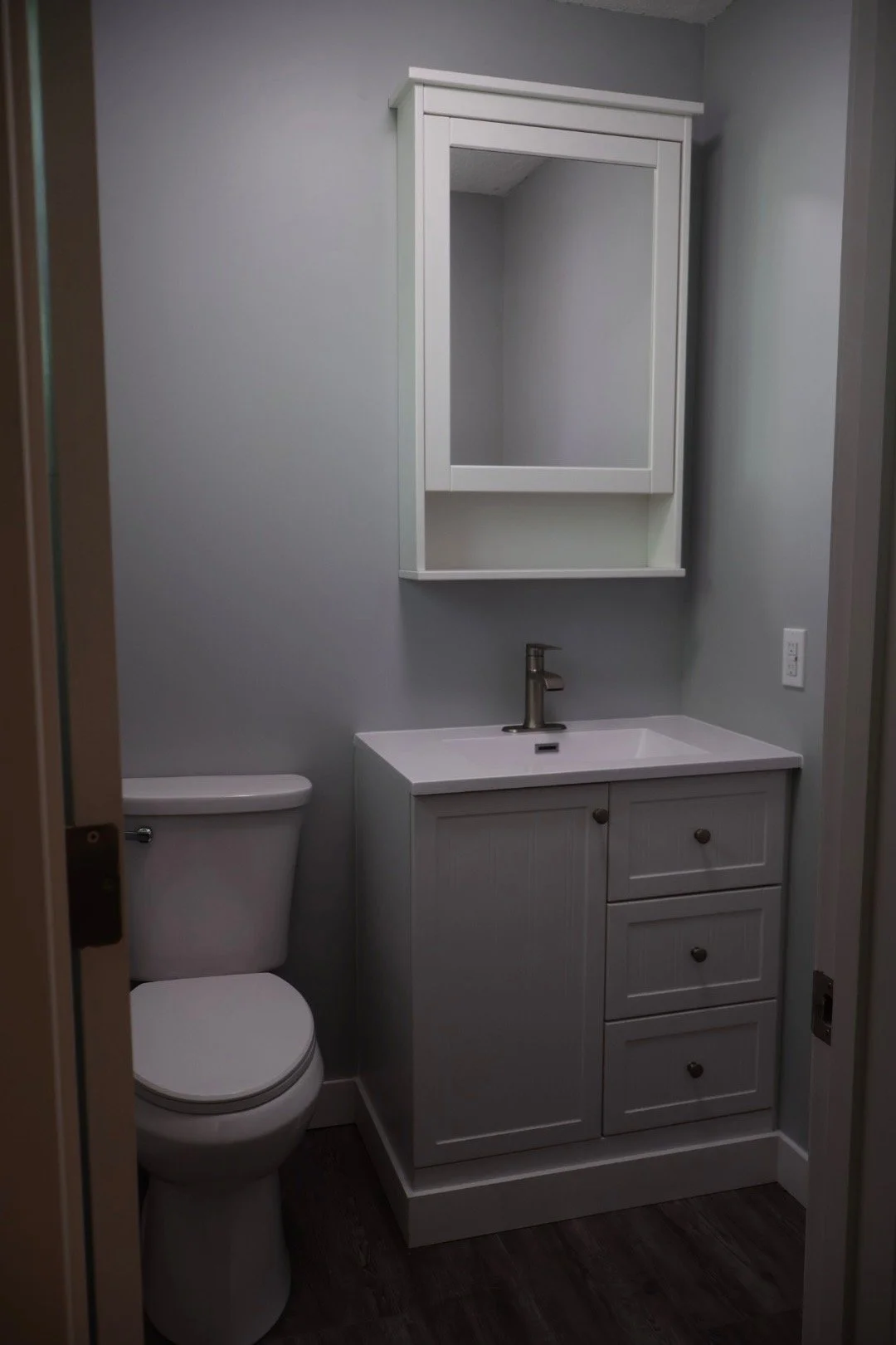 A small bathroom with a white toilet, a white vanity with a sink, a gray wall, a mirror cabinet, and a dark wood floor.