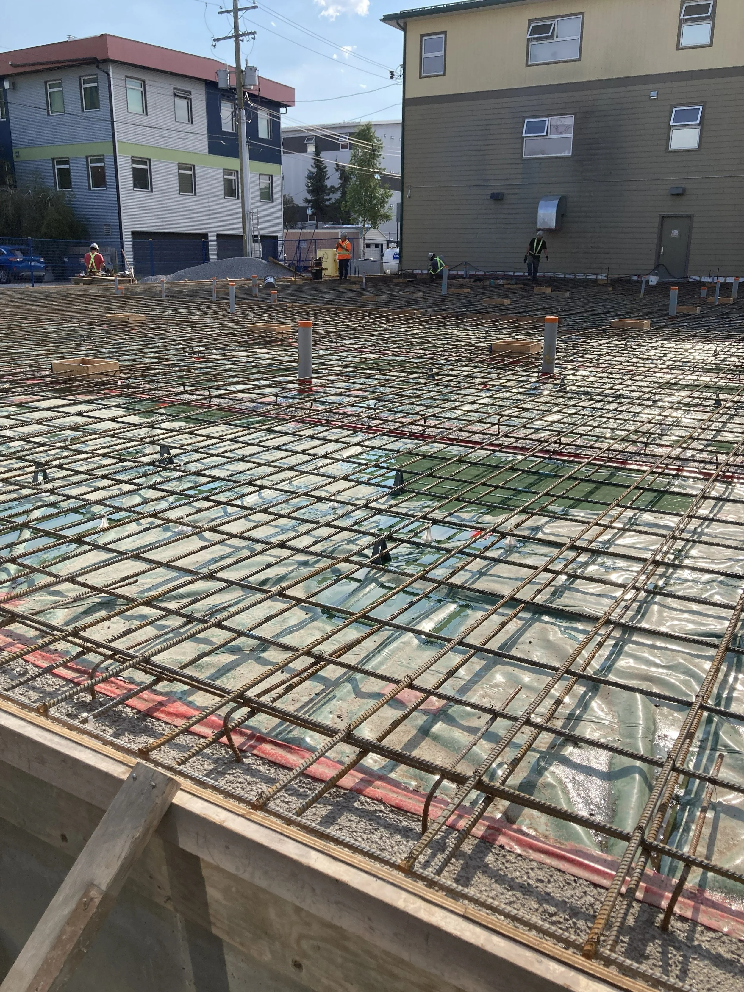 Construction site with rebar grid and workers wearing safety vests and helmets, preparing for concrete pouring near residential buildings.