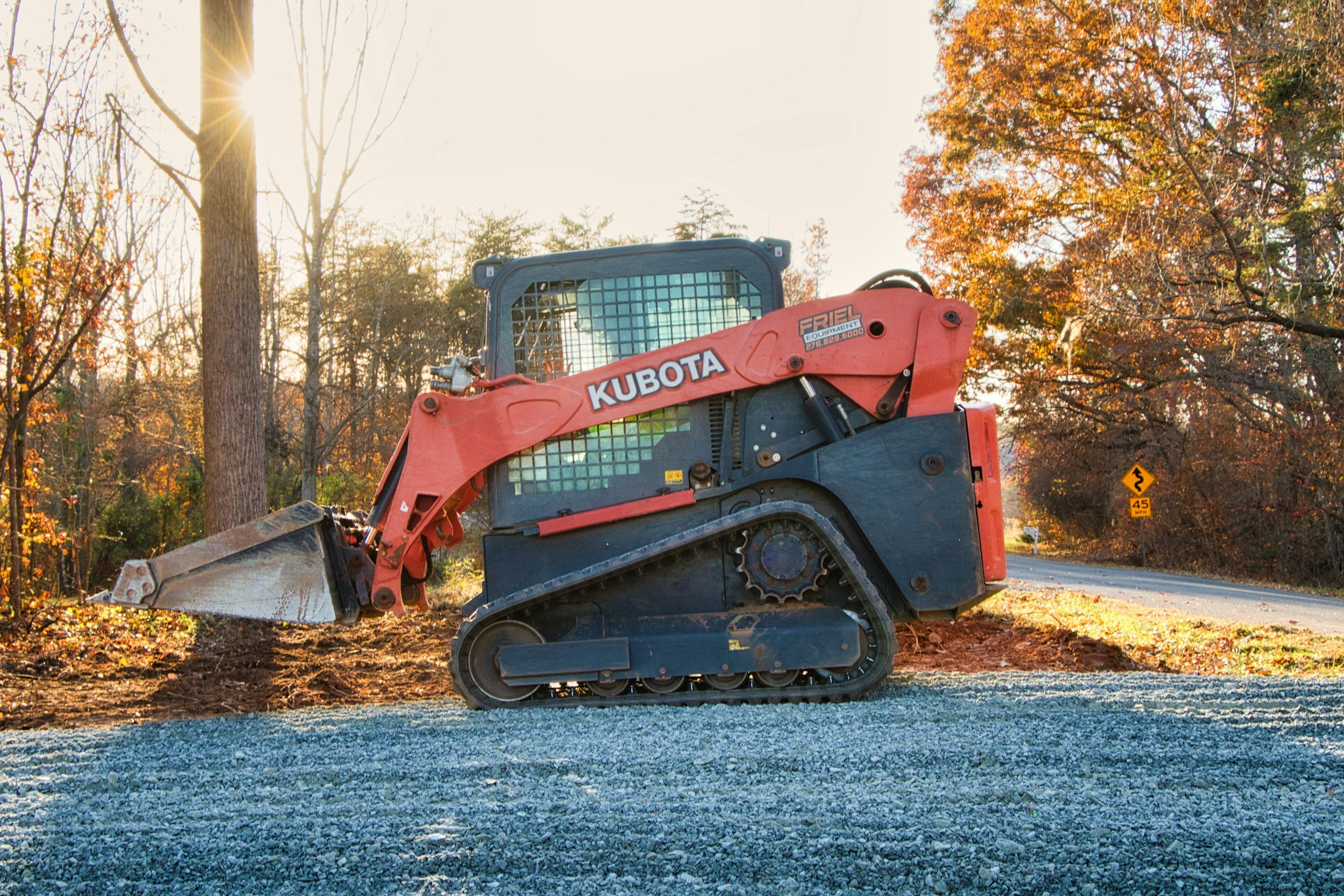 A small Kubota track loader with orange and black body is working on a gravel surface, surrounded by trees with fall foliage and a bright sun in the background.