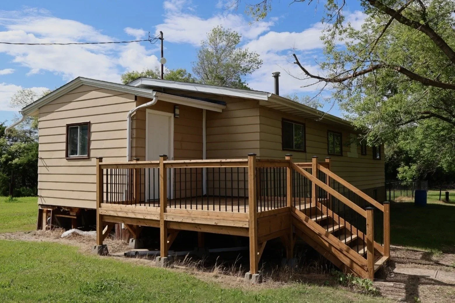 A beige house with a small wooden porch, stairs, and a grassy yard, with trees and a blue sky in the background.
