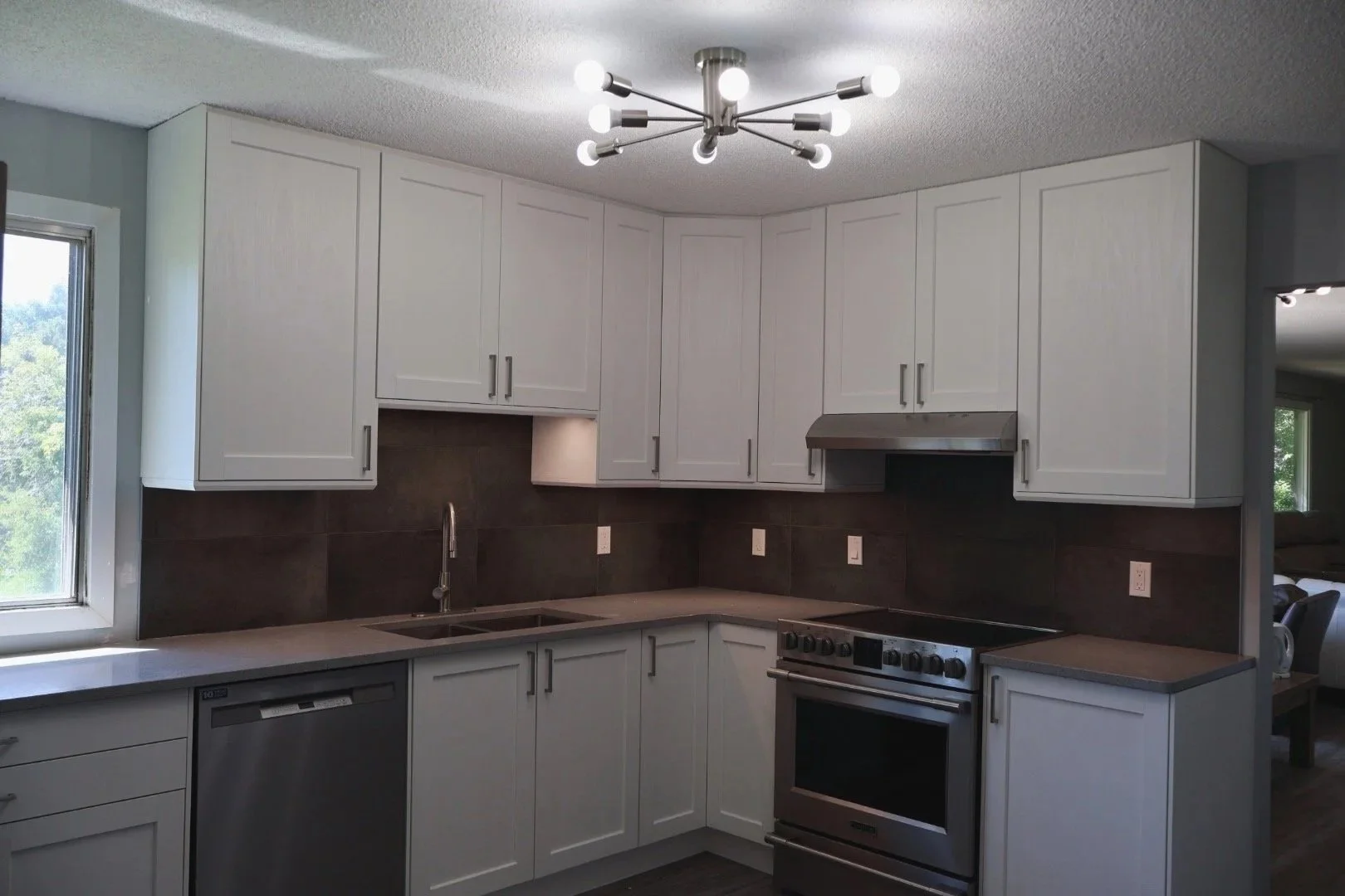 Modern kitchen with white cabinets, black backsplash, stainless steel appliances, window with outdoor view, and geometric ceiling light fixture.