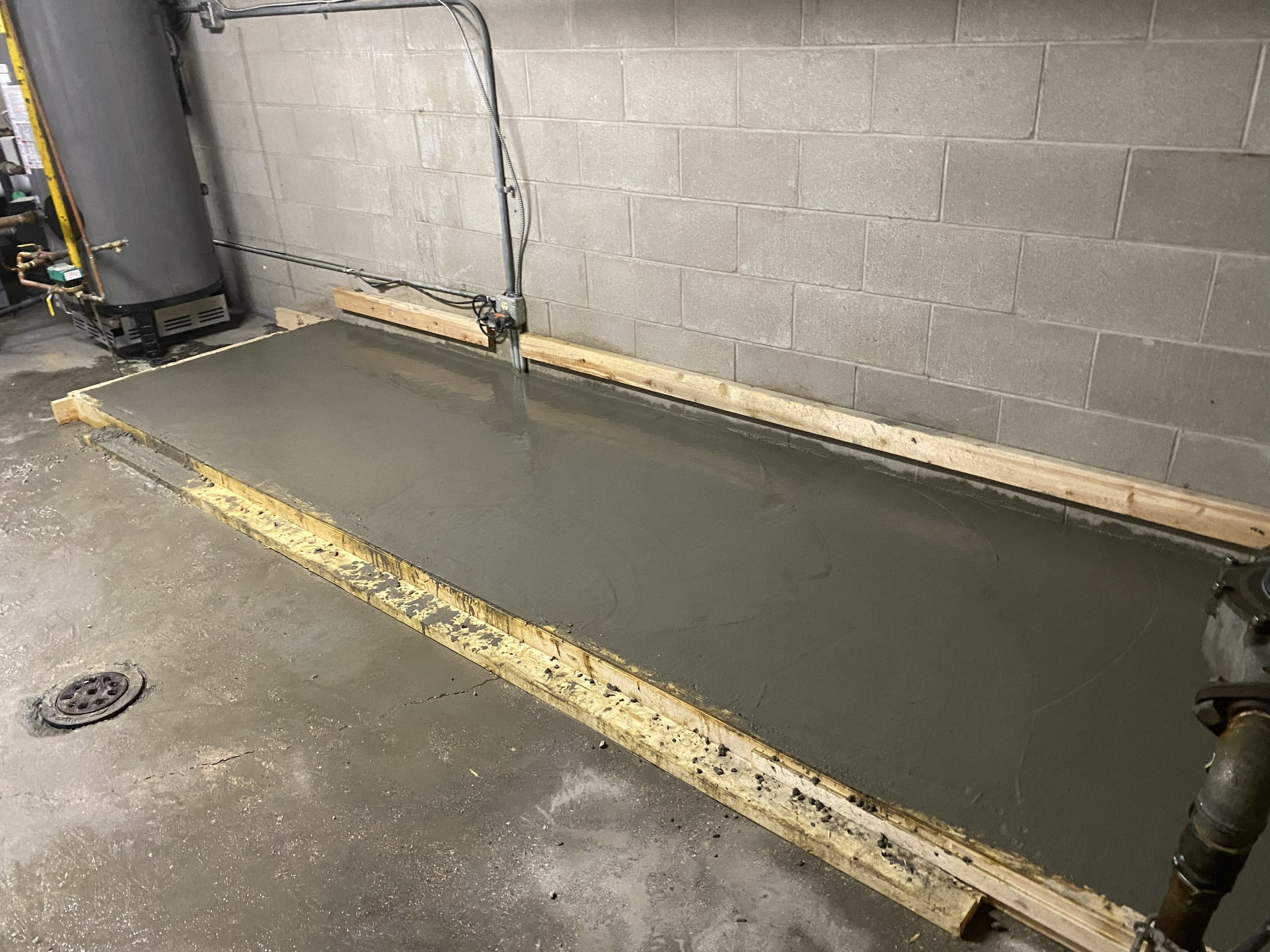 Concrete slab leveled in a wooden frame against a cinder block wall with electrical conduit and wiring.