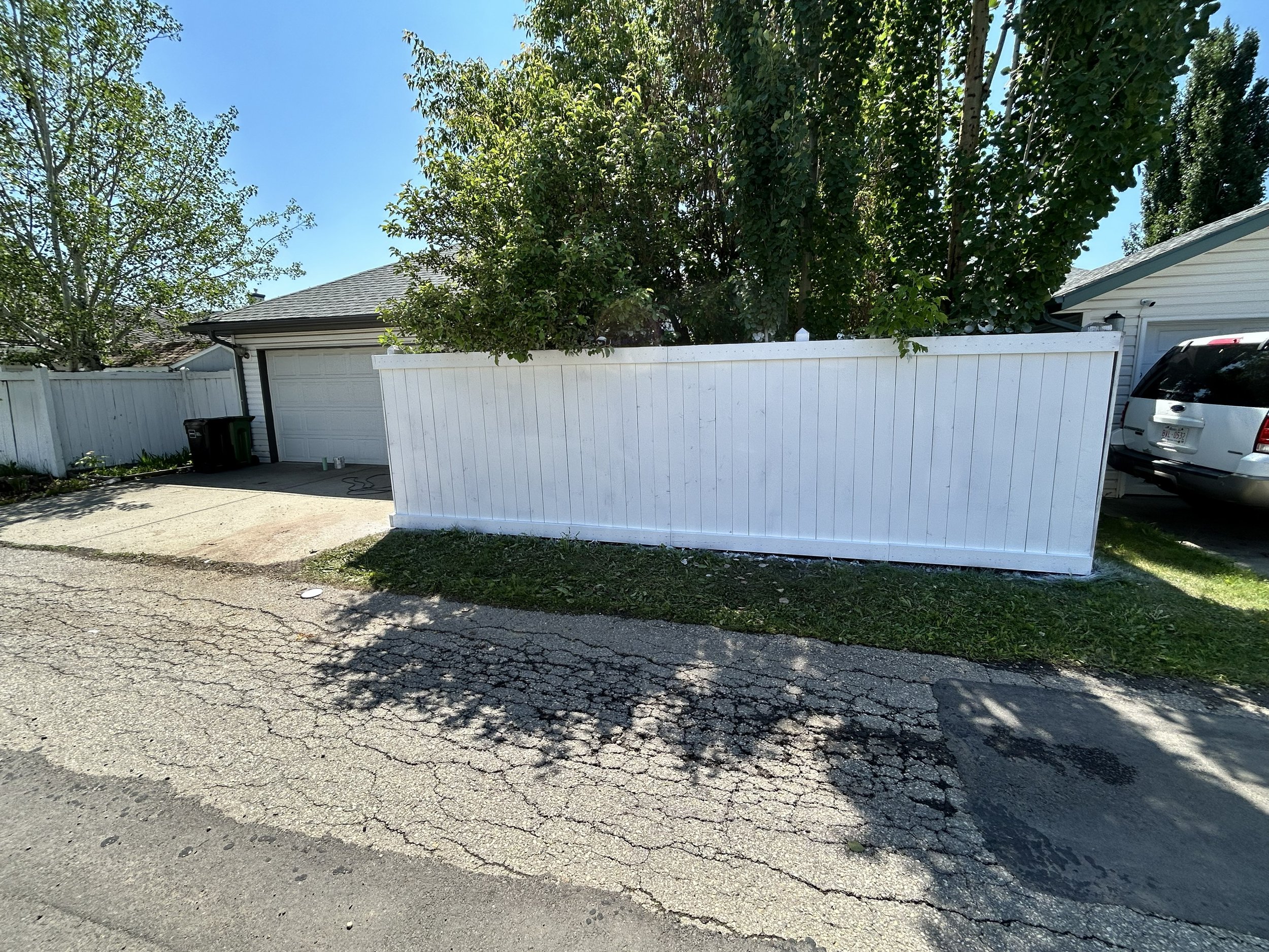 A residential driveway with a white fence, surrounded by trees, a gray house, a car, and a green trash bin, under a clear blue sky.