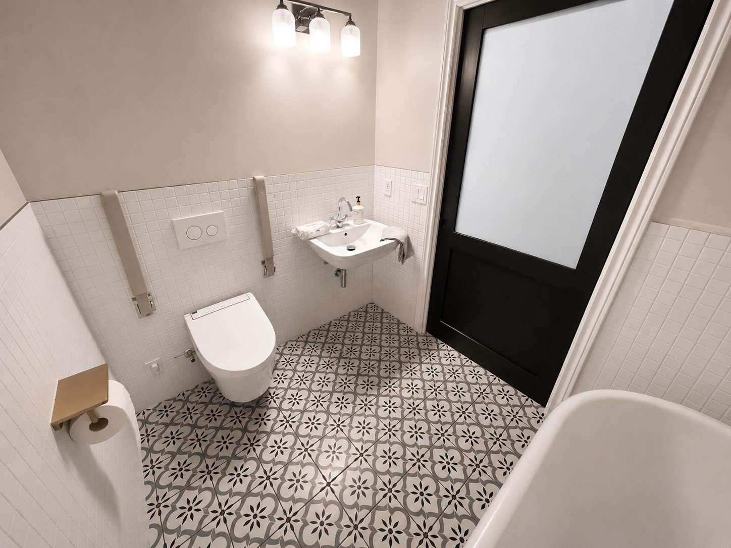 Small bathroom with a wall-mounted toilet, a tiny sink, a frosted glass door, patterned floor tiles, and a three-light ceiling fixture.