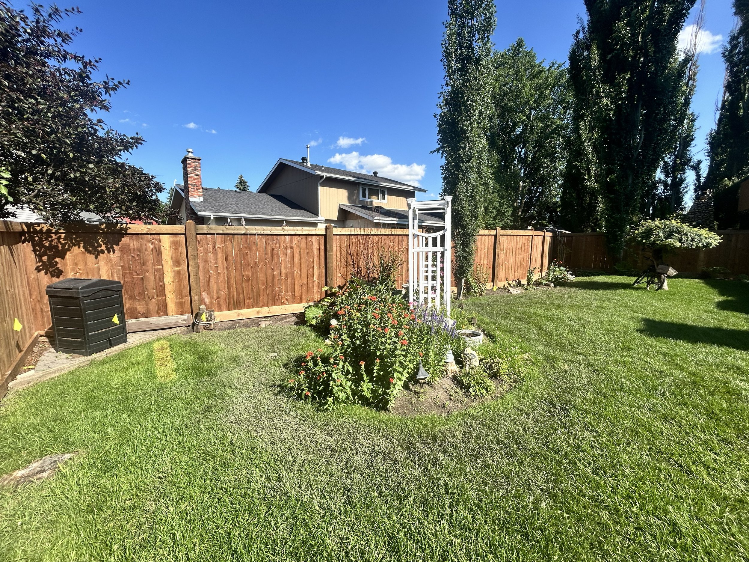 A backyard with a wooden fence, a garden with flowering plants, a grassy lawn, a large tree, and a house in the background under a clear blue sky.