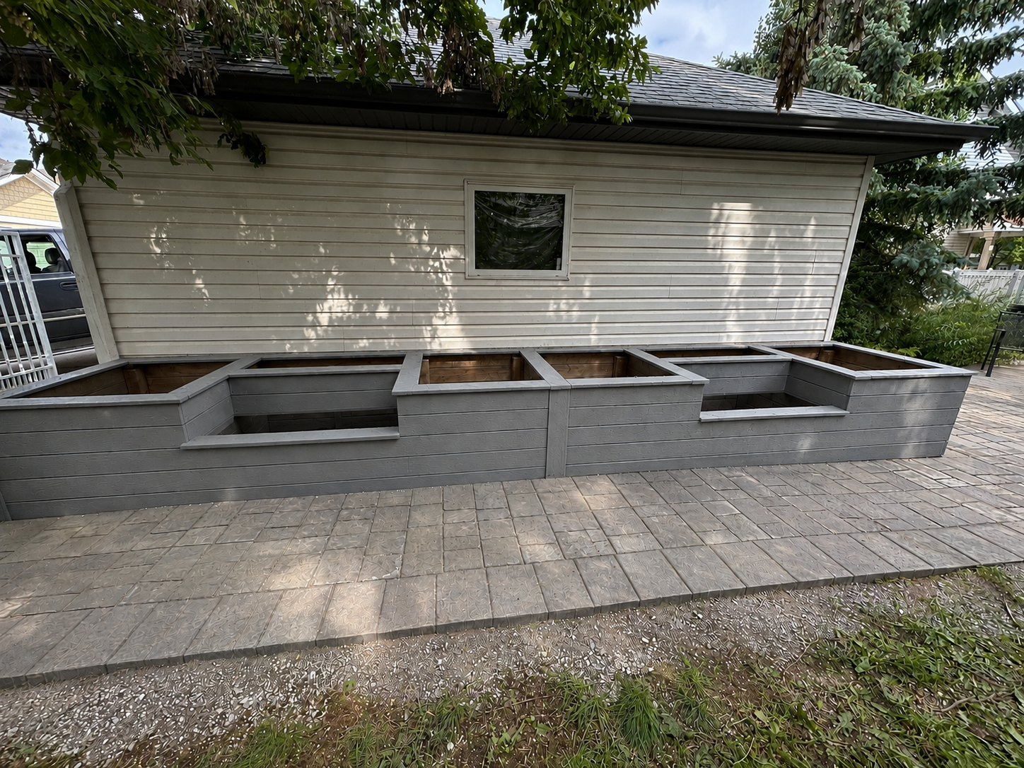 Backyard vegetable garden beds under construction, with empty wooden frames on a concrete patio next to a house with white siding and a small window.