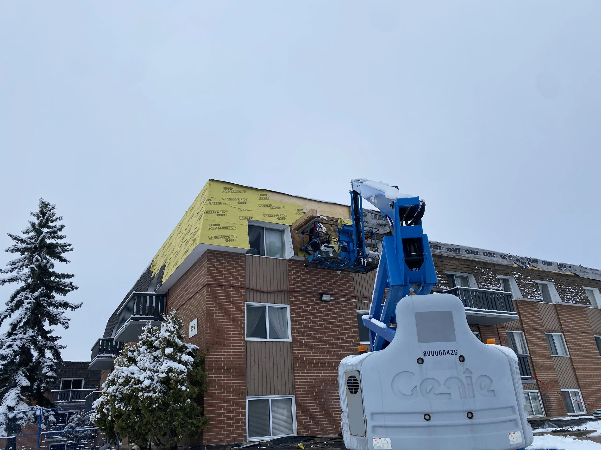 A construction worker using a boom lift to work on the upper part of a residential building with a brick exterior and a partially covered roof, surrounded by snowy trees on a cloudy day.