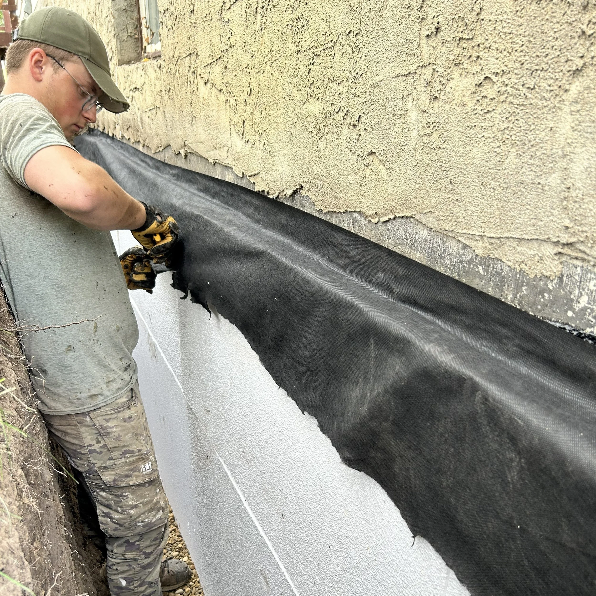 A person wearing glasses, a cap, and camouflage pants is working on a building exterior, applying or repairing a black waterproofing membrane on the foundation wall.