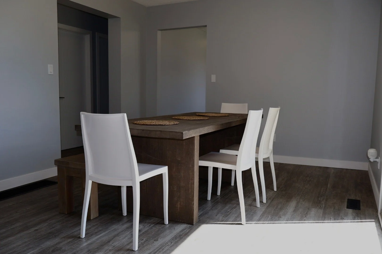 A dining room with a wooden table and four white chairs, placed on a wooden floor against a gray wall.