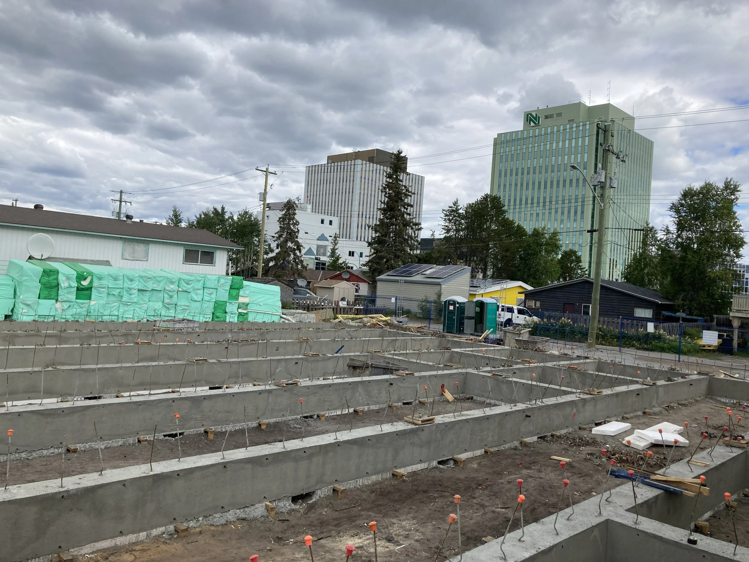 Construction site with concrete foundation and reinforcement bars in foreground, residential buildings with solar panels and taller office buildings in background, cloudy sky overhead.