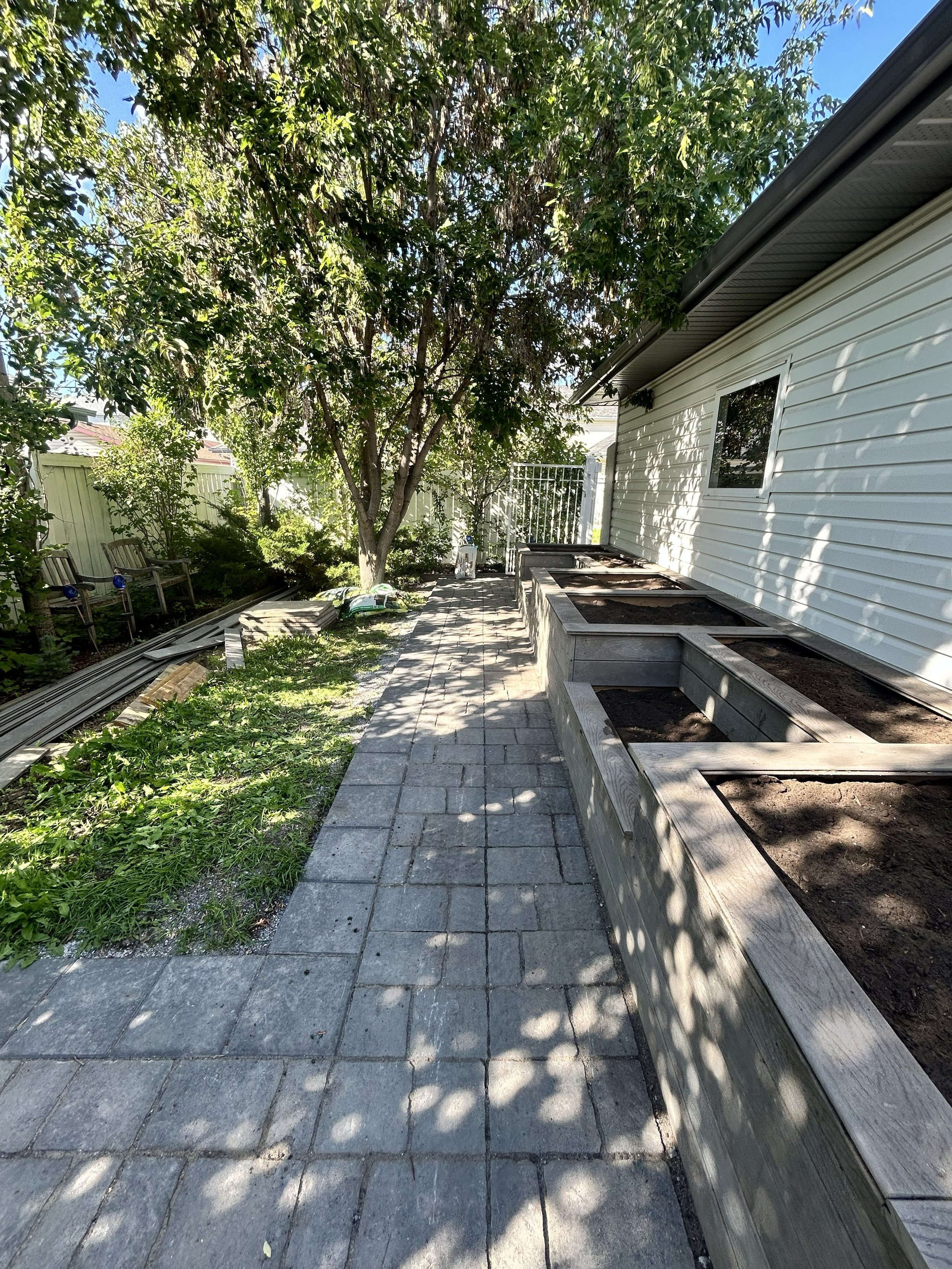 A backyard patio with paver stones, a white house wall on the right, and a garden bed with soil in wooden frames, a tree with green leaves casting shadows, and garden furniture to the left, under a clear blue sky.