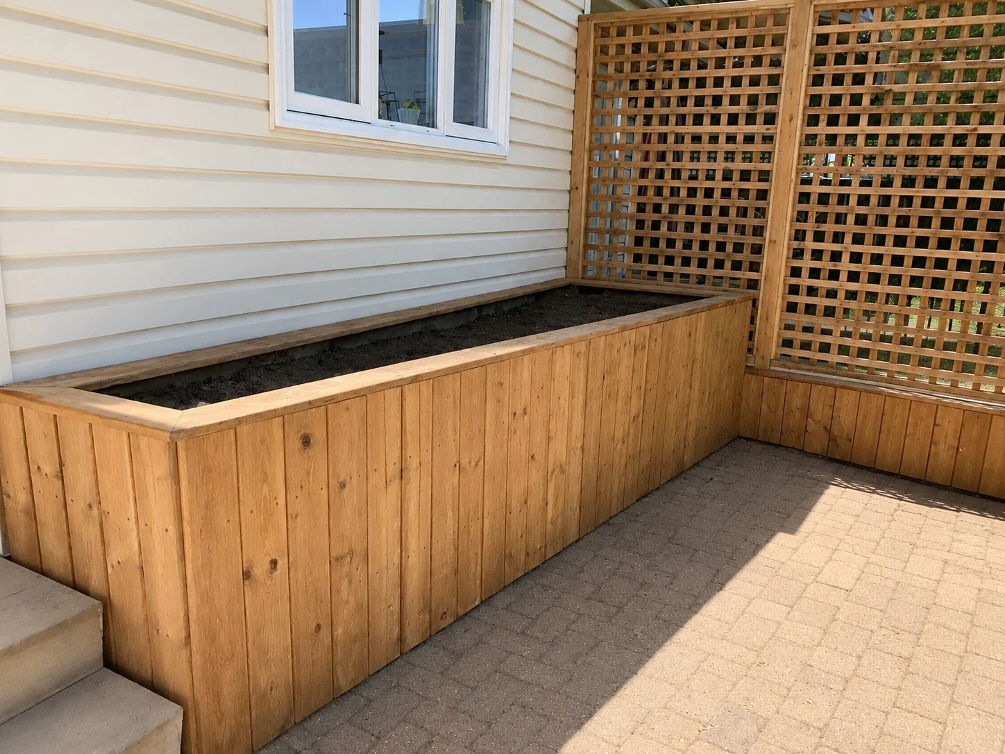 Empty wooden garden bed with soil, situated beside a house with white siding and a lattice fence on the right side.