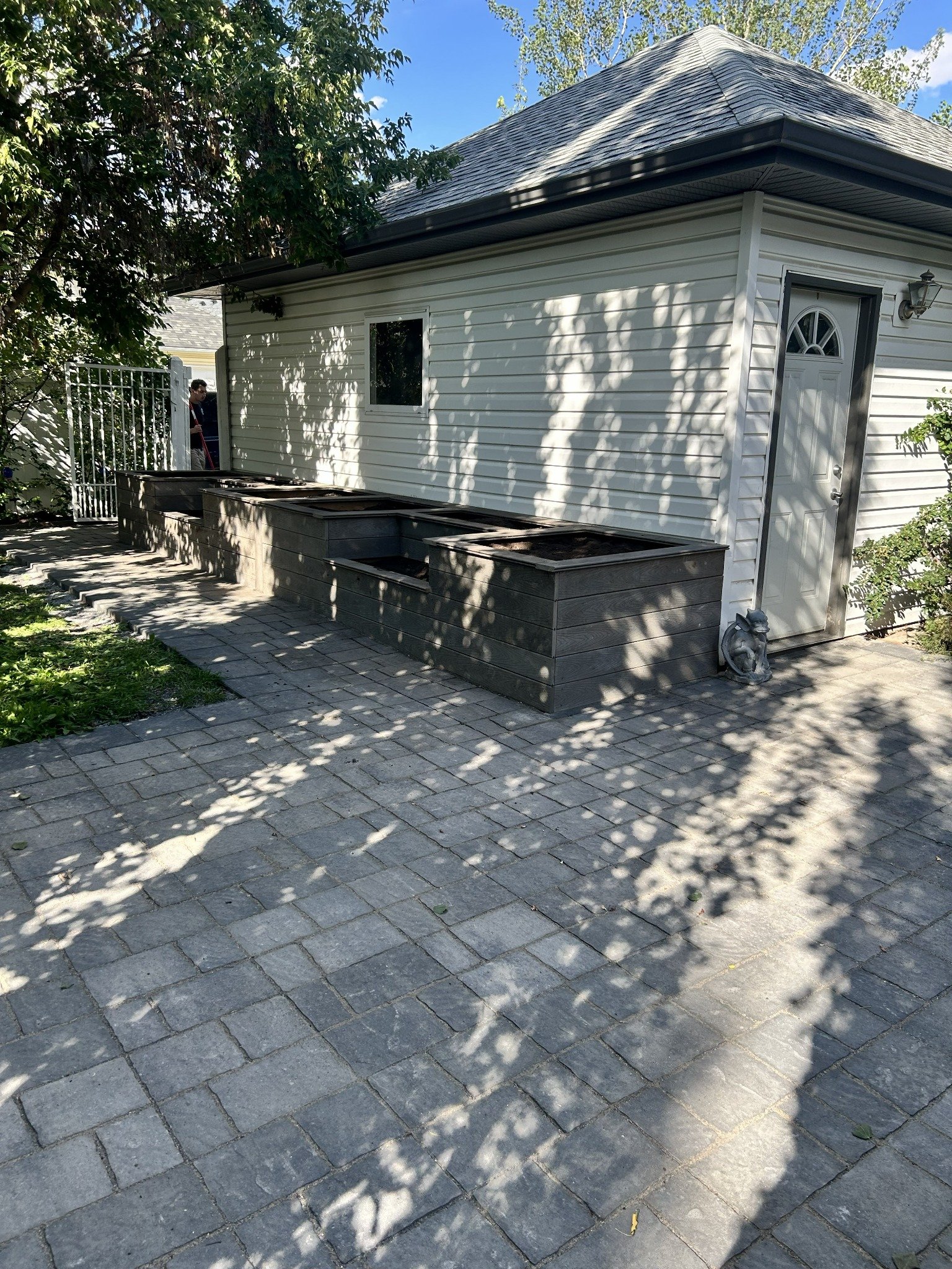 Backyard with a white house, a small window, a door with a semi-circular window, and metal planters on a stone patio. Shadows of leaves cast on the house and patio.