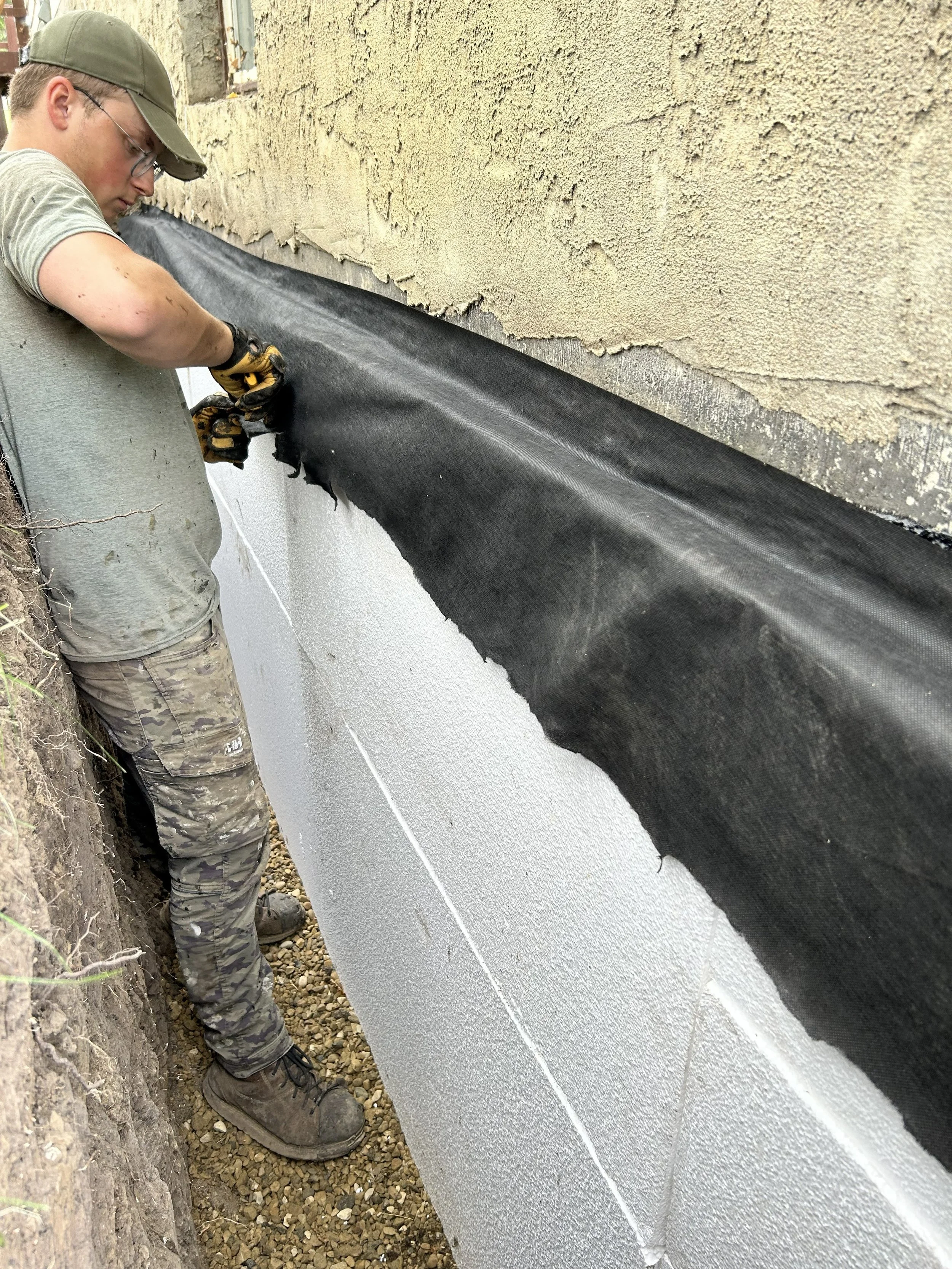 A person wearing glasses, a green cap, and camouflage pants working on the exterior wall of a building, installing a black waterproofing membrane along the base of the wall.