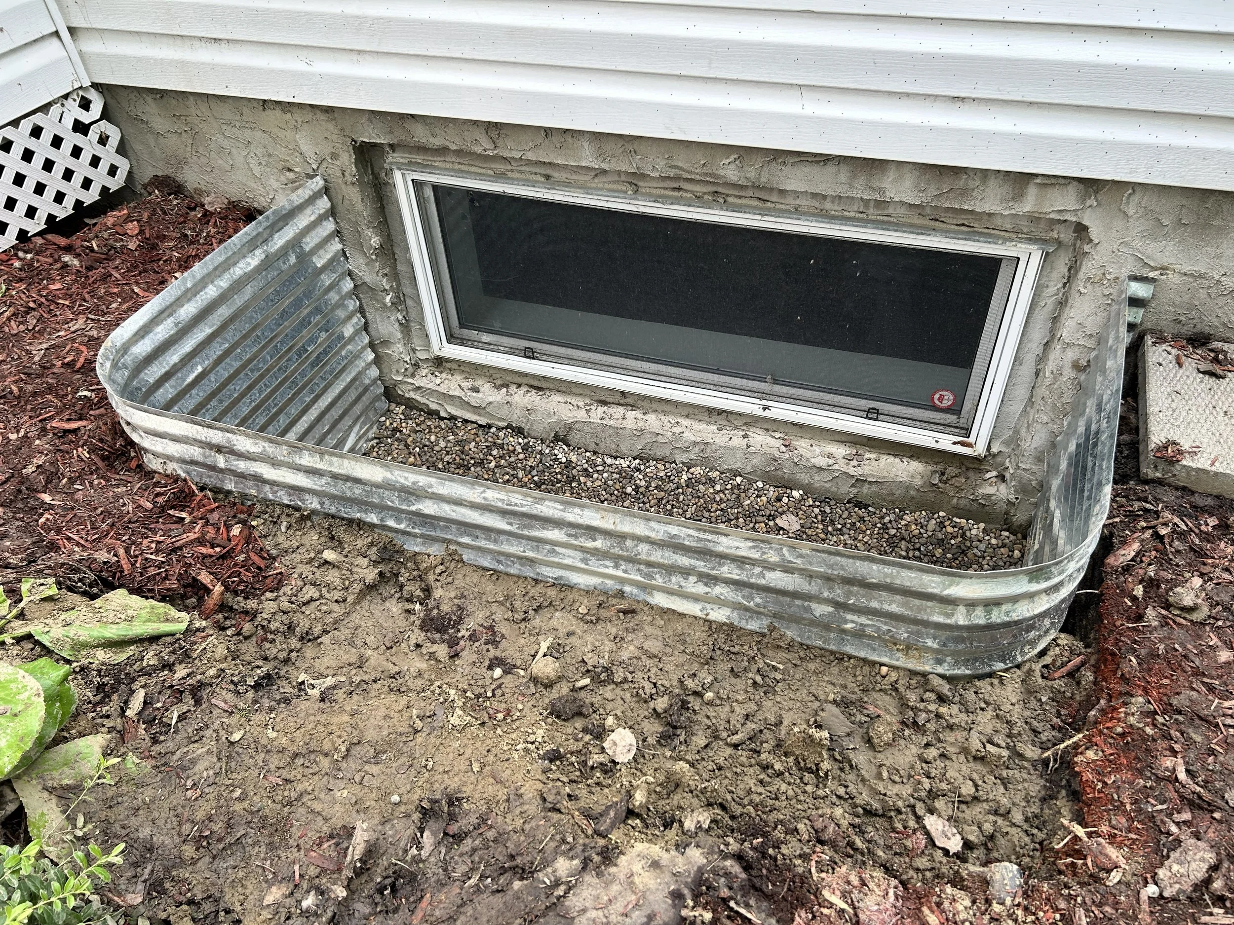 The image shows a basement window installed in the foundation wall, surrounded by rough concrete. There are metal window well supports on each side, with gravel at the bottom inside the metal supports, and dirt and mulch outside around the window.
