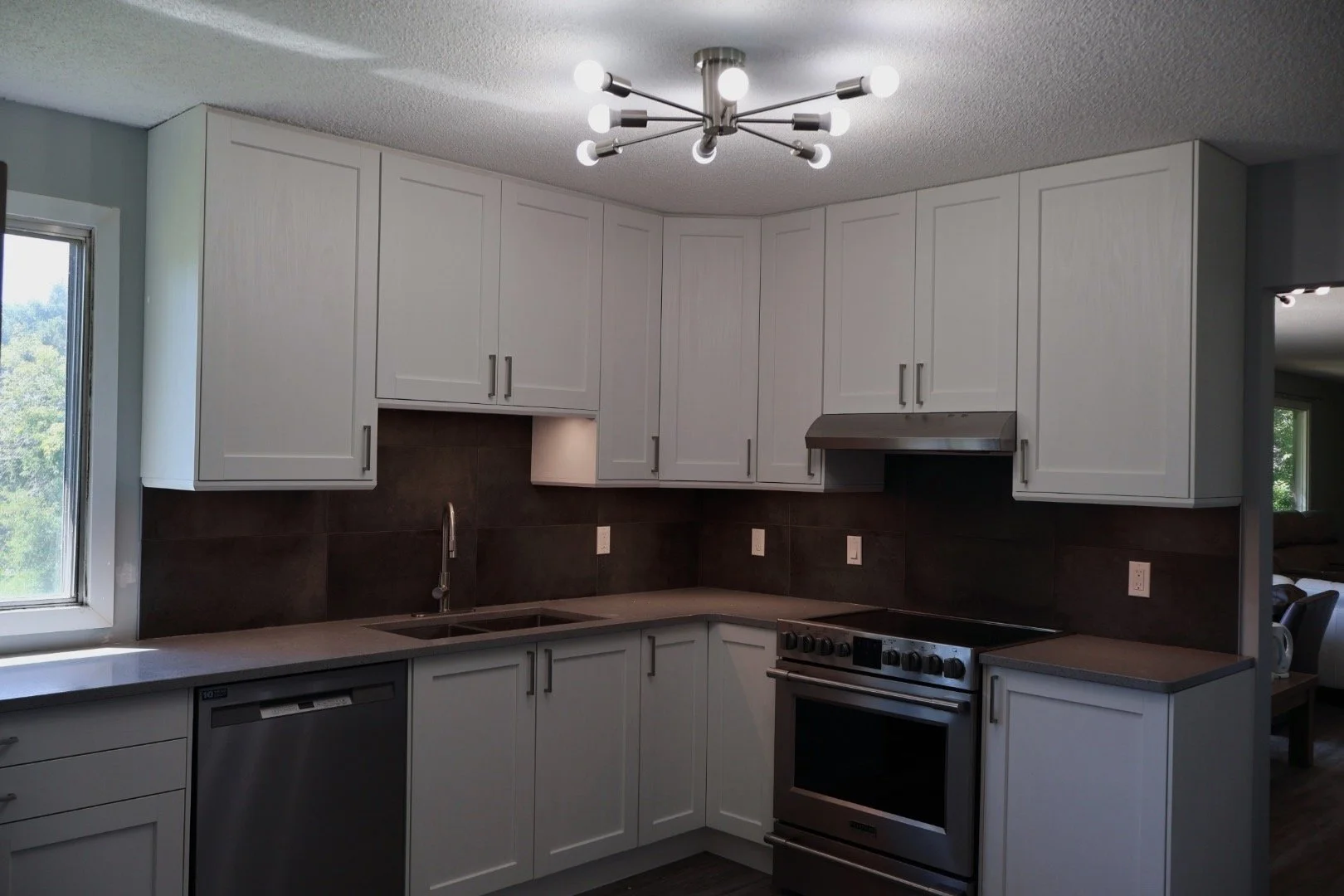 Modern kitchen with white cabinets, black backsplash, stainless steel stove, and dishwasher, illuminated by a ceiling chandelier and natural light from a nearby window.