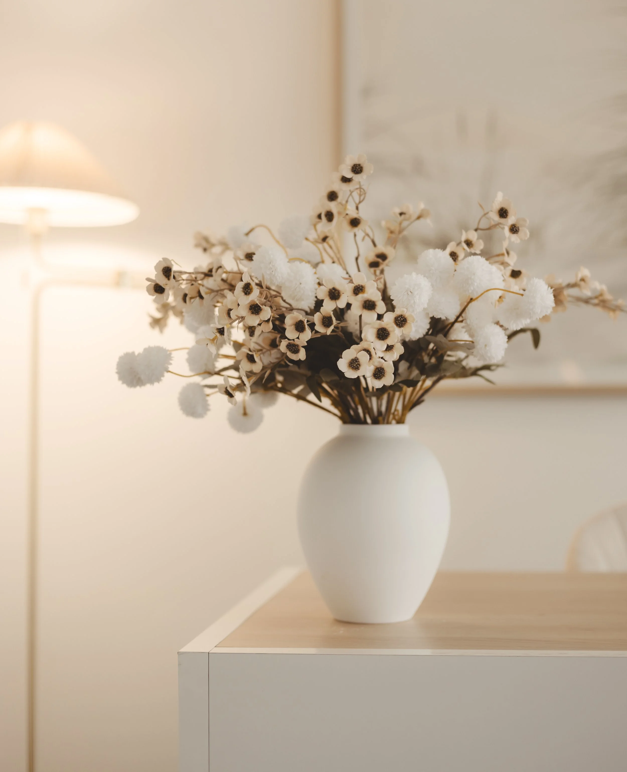 A white vase with a bouquet of white and brown flowers on a wooden table in a softly lit room.