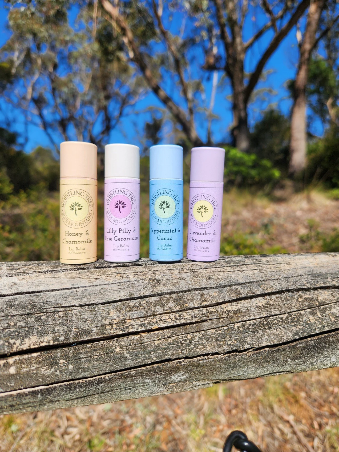 Four cardboard lip balm tubes are displayed on a weathered wooden surface outdoors with trees and a blue sky in the background. The flavors are Honey & Chamomile, Lilly Pilly & Rose Geranium, Peppermint & Cacao, and Lavender & Chamomile.