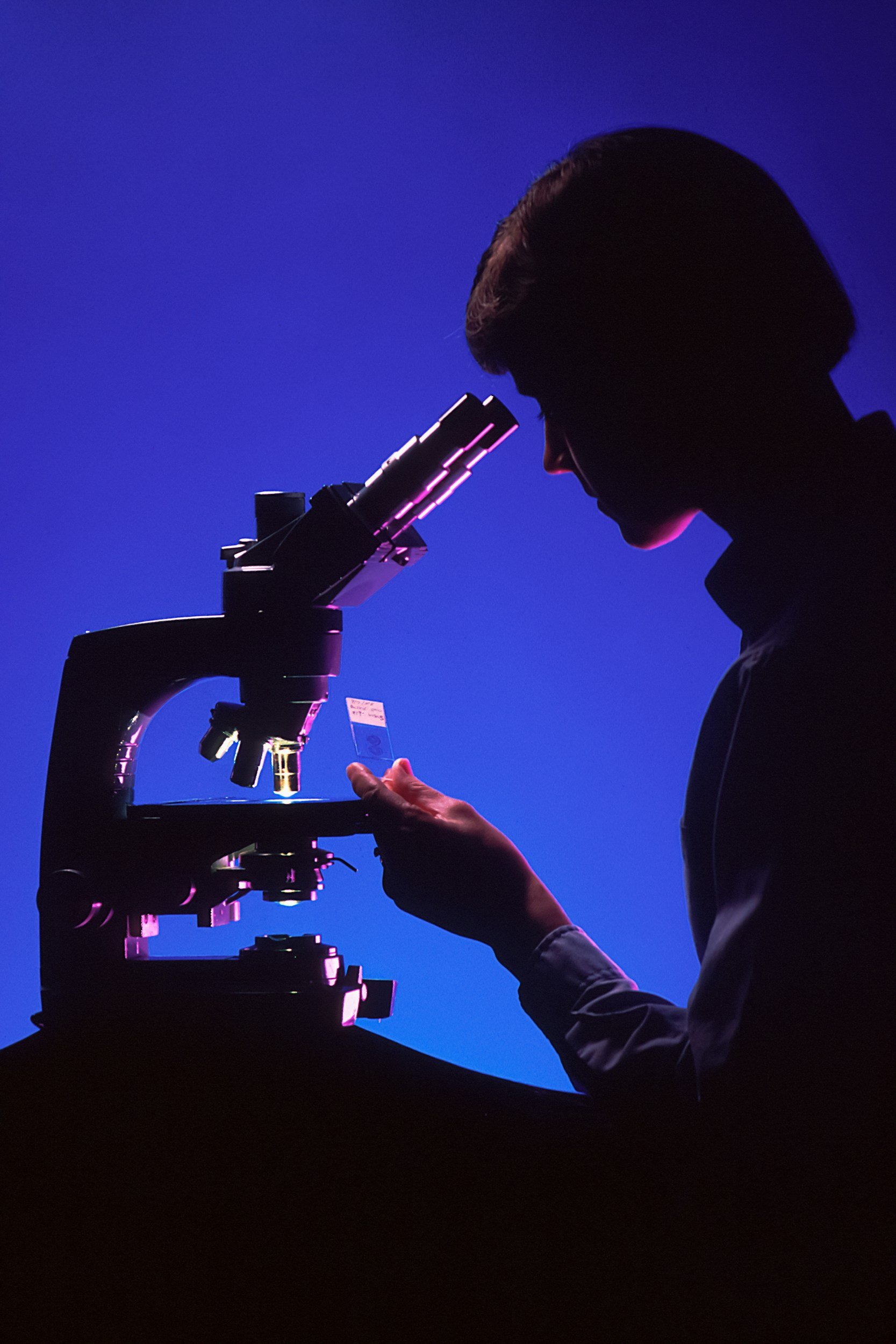 Silhouette of a scientist examining a slide under a microscope against a blue background.