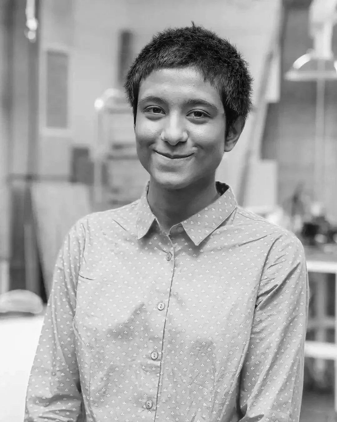 A young person with short hair smiling in a casual patterned button-up shirt, standing indoors with a blurred background.